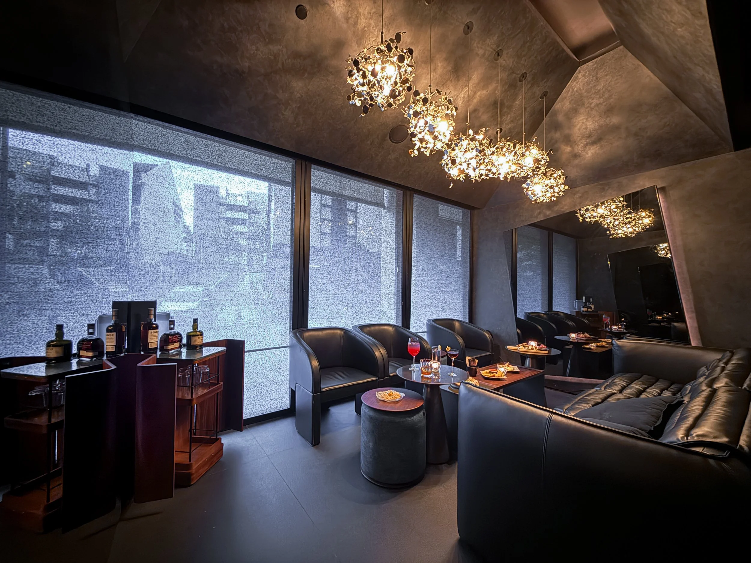 Luxurious private room with leather chairs and a sofa, round tables with drinks, modern chandelier lighting, large tinted windows, and a wall-mounted TV.
