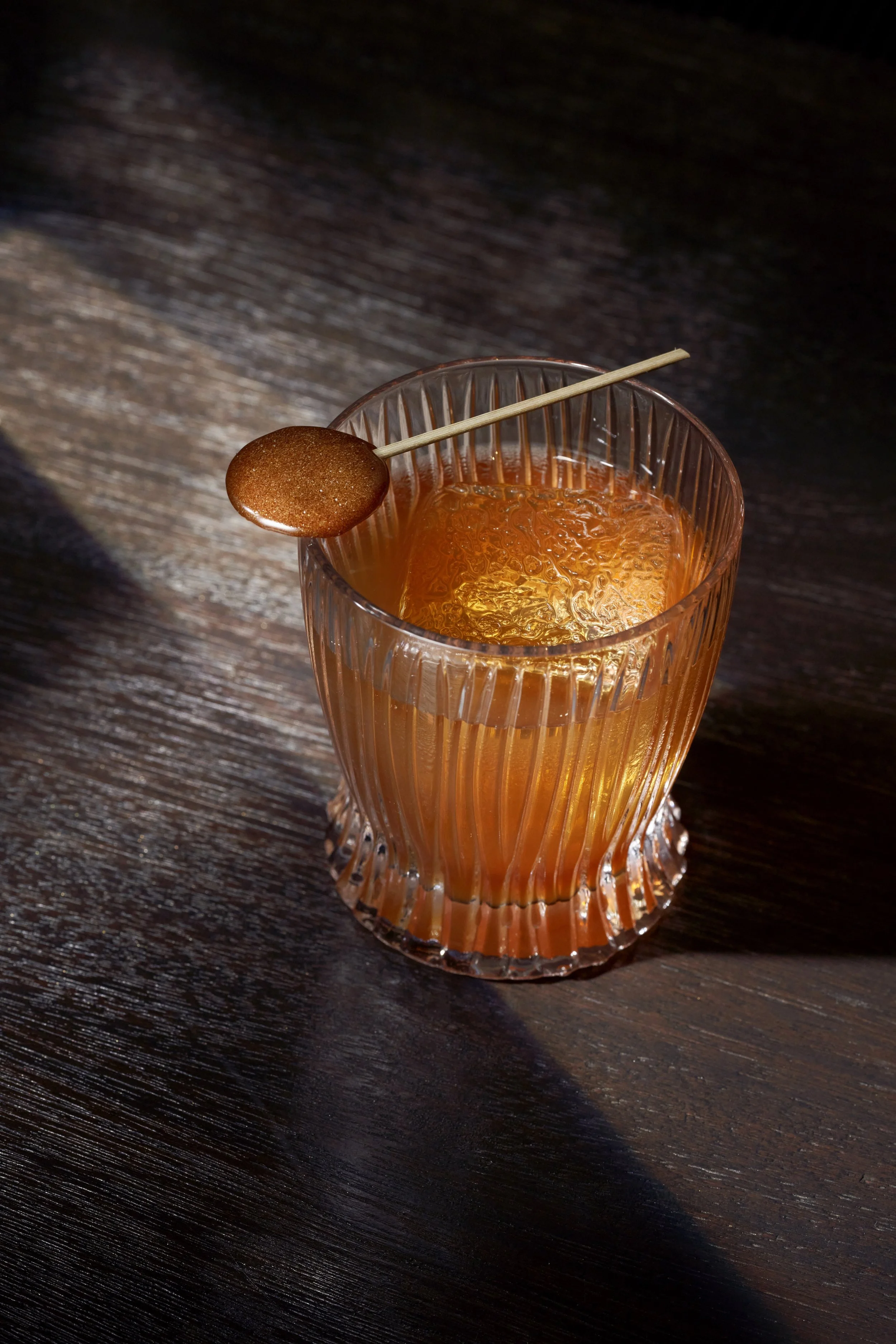 A classic cocktail like an old fashioned with a brown lollipop on a skewer resting on top of the glass as a garnish, placed on a dark wooden surface.