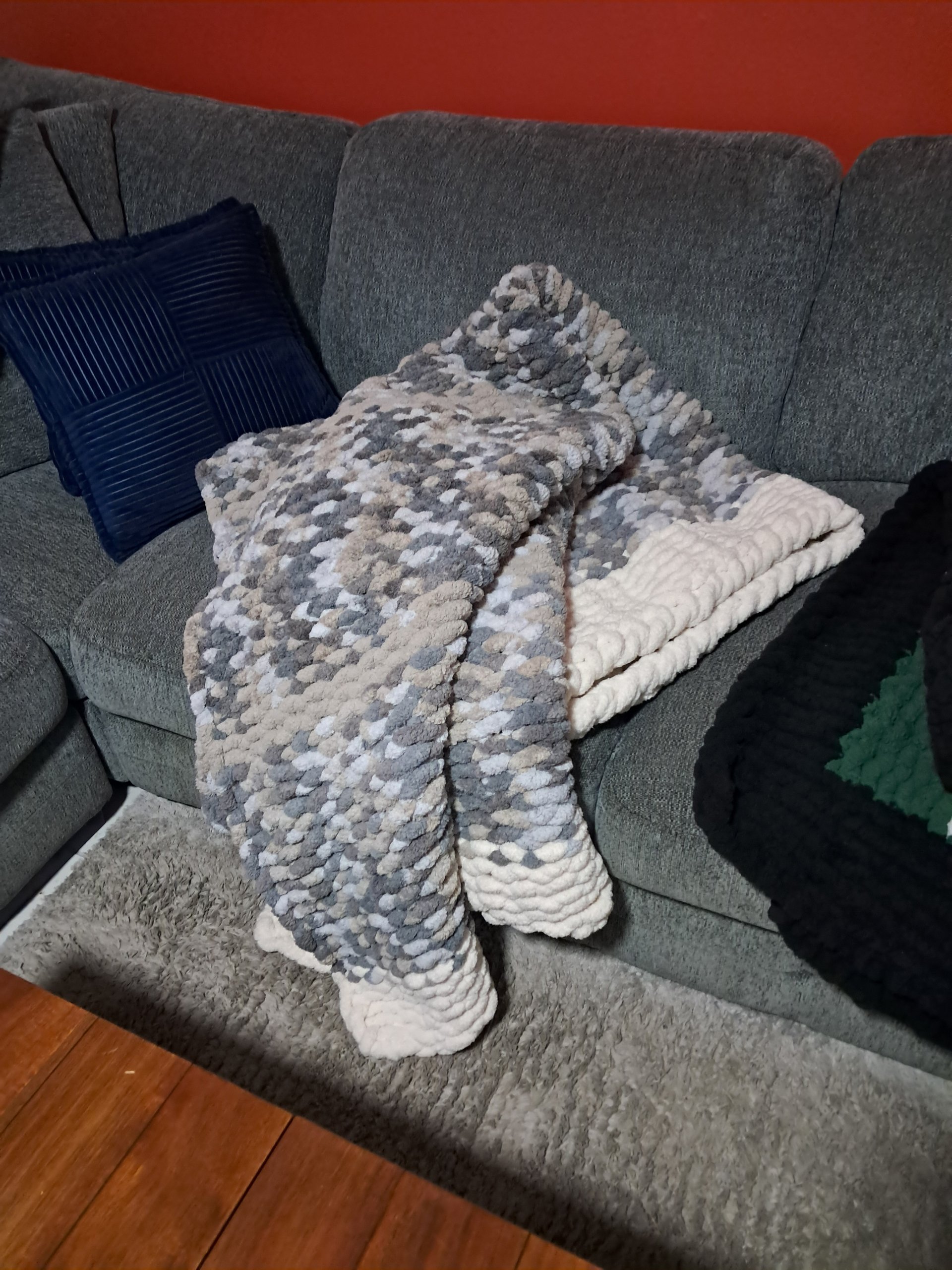 A grey sectional sofa with a navy blue pillow, layered with a chunky grey and white blanket and a matching white blanket, with a black and green blanket on the right side. A reddish-orange wall is in the background, and a light grey textured rug is on the wooden floor in front of the sofa.