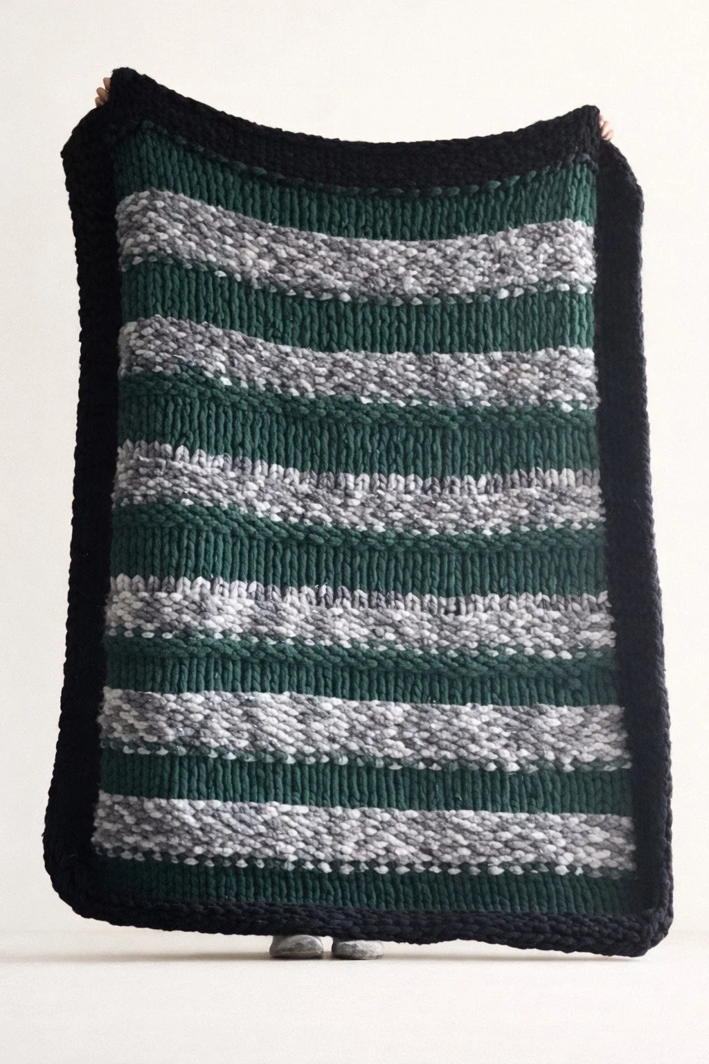 A rectangular, striped, handwoven rug with green, gray, and black stripes, standing upright on a white surface against a plain white background.