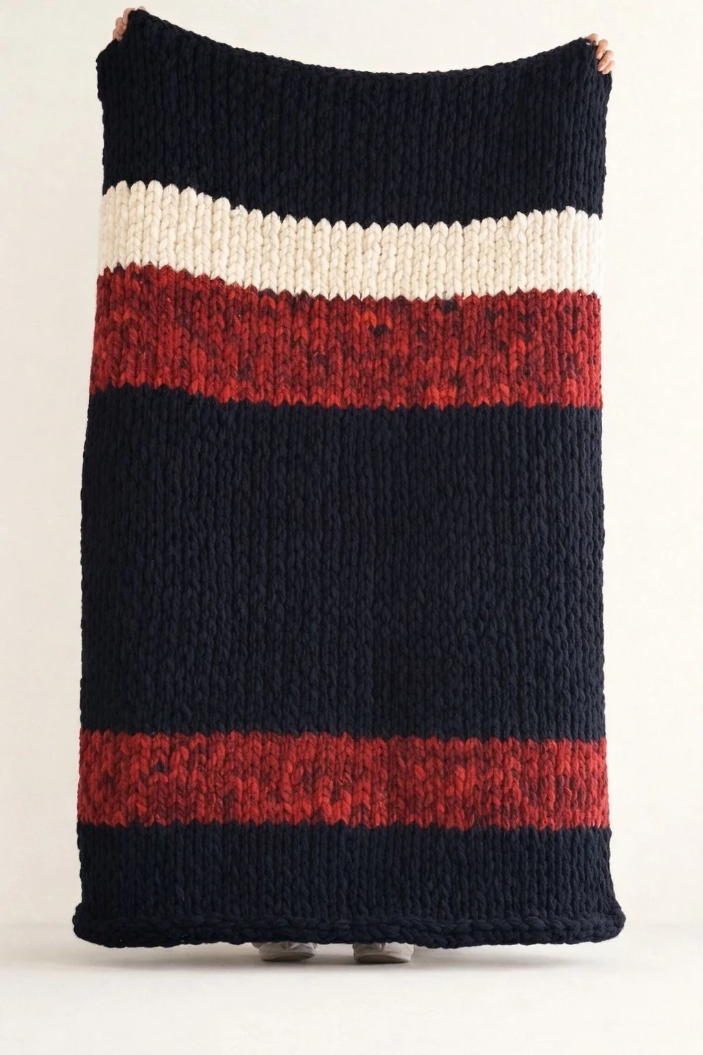 Person holding a large, striped, knitted blanket with navy, white, red, and navy stripes against a plain background.