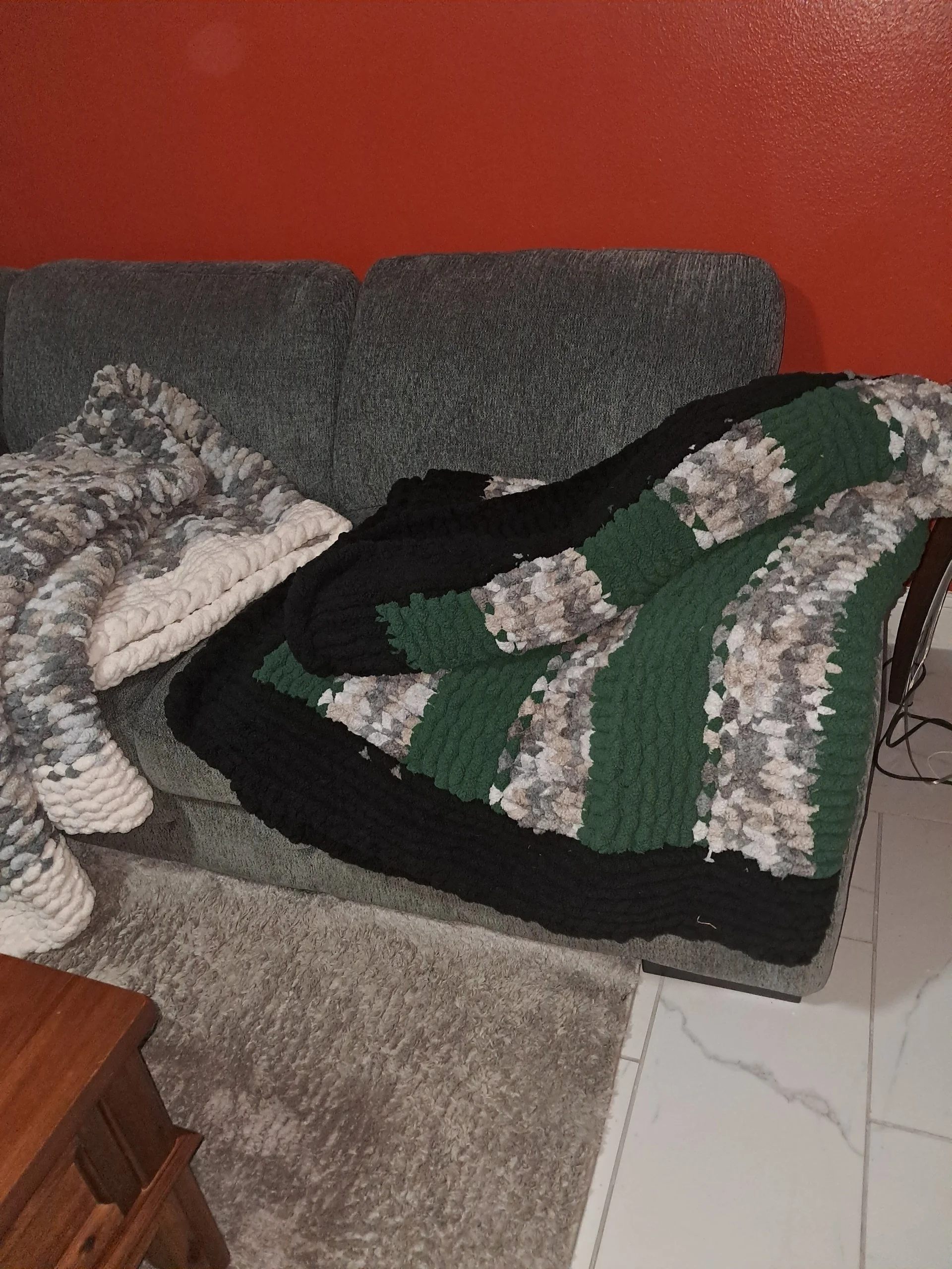 A gray sofa with two folded chunky knit blankets, one in black and one with a multicolored pattern, draped over it, in a room with an orange wall and a gray area rug.