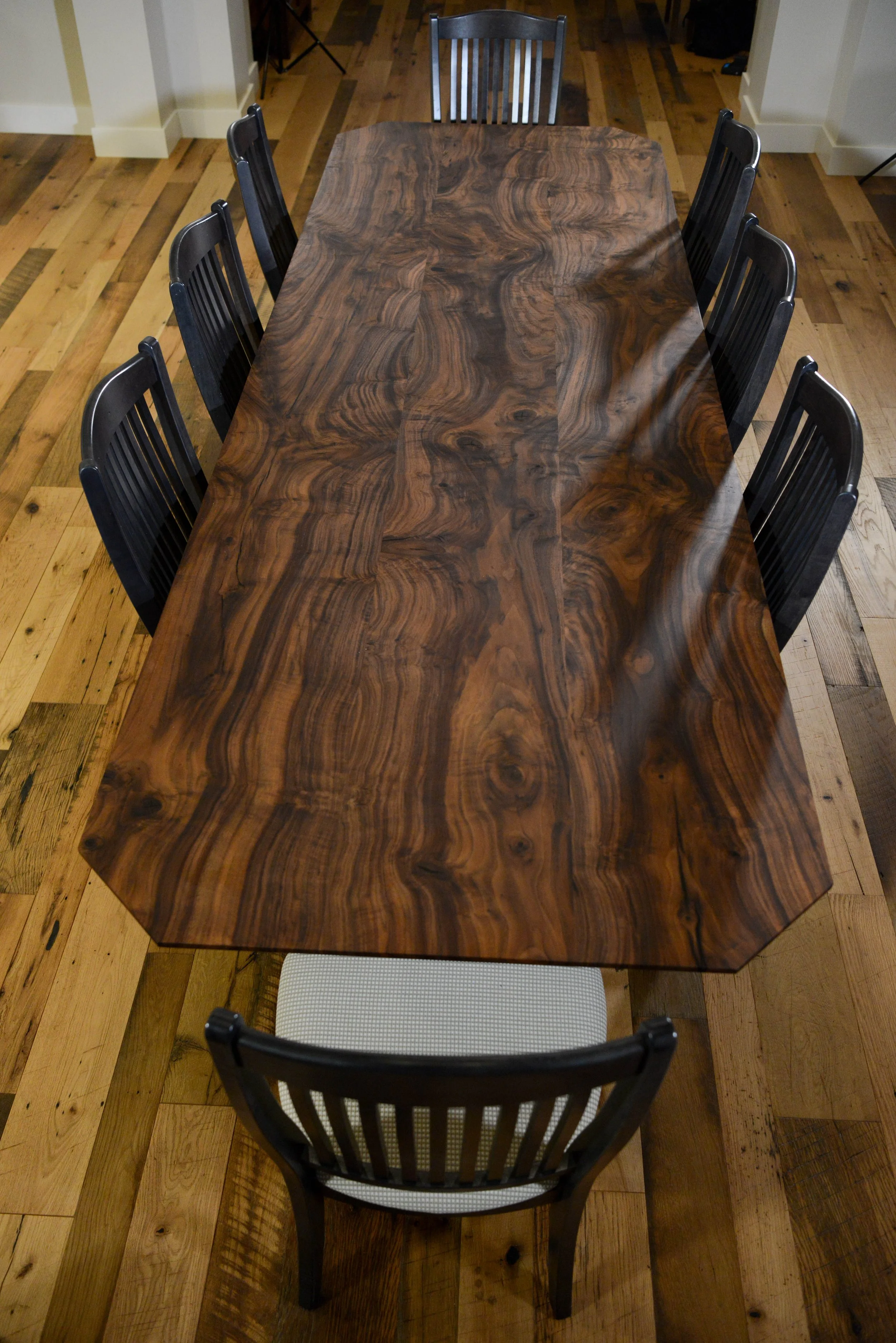 Labrado Forge Blacksmithing A large wooden dining table with a polished surface and a natural wood grain pattern, surrounded by eight chairs, six black and one white, in a room with hardwood floors.