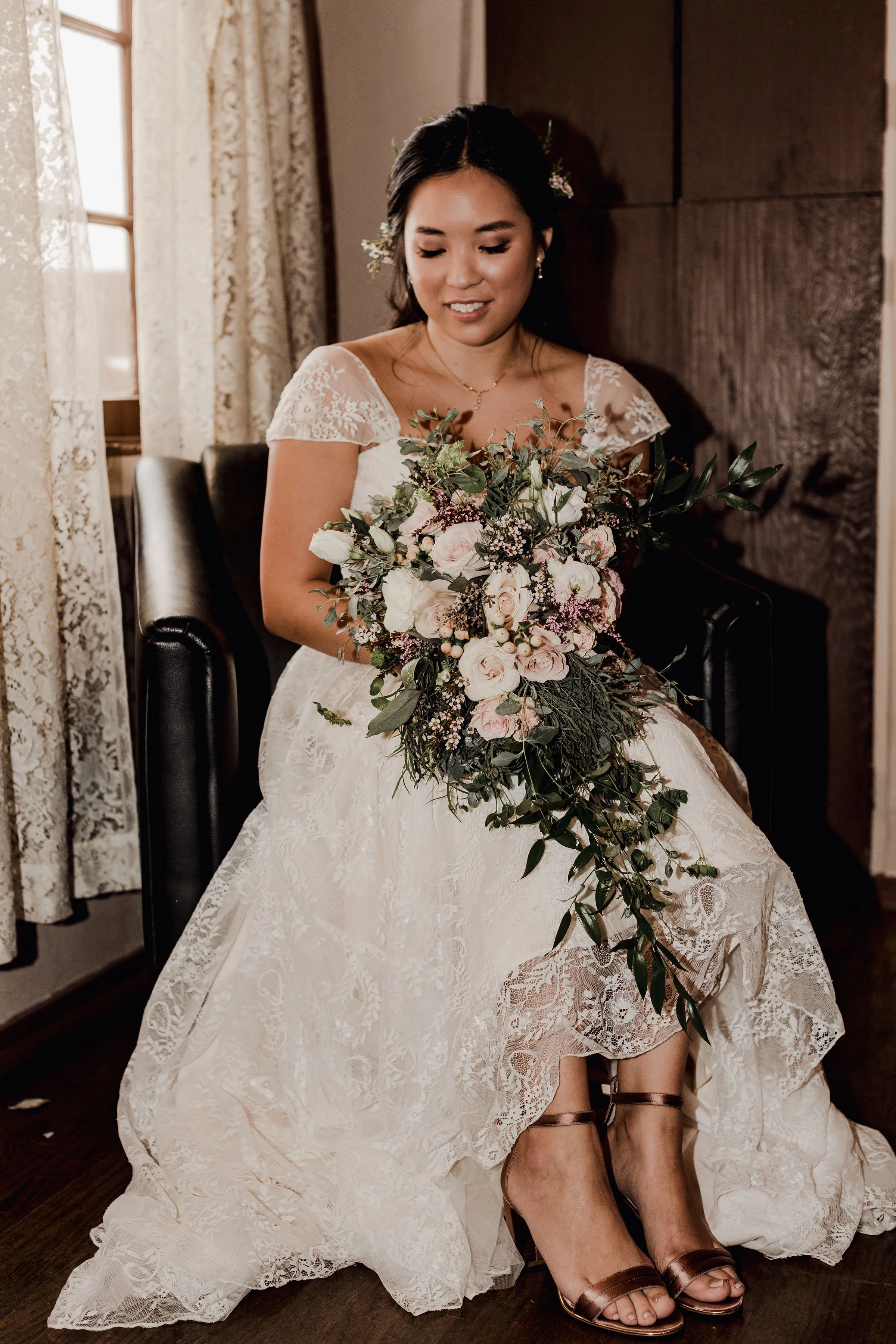 A bride sitting down in a white lace wedding dress, holding a large bouquet of pink and white flowers with greenery, smiling softly, with her feet in bronze high heel sandals.