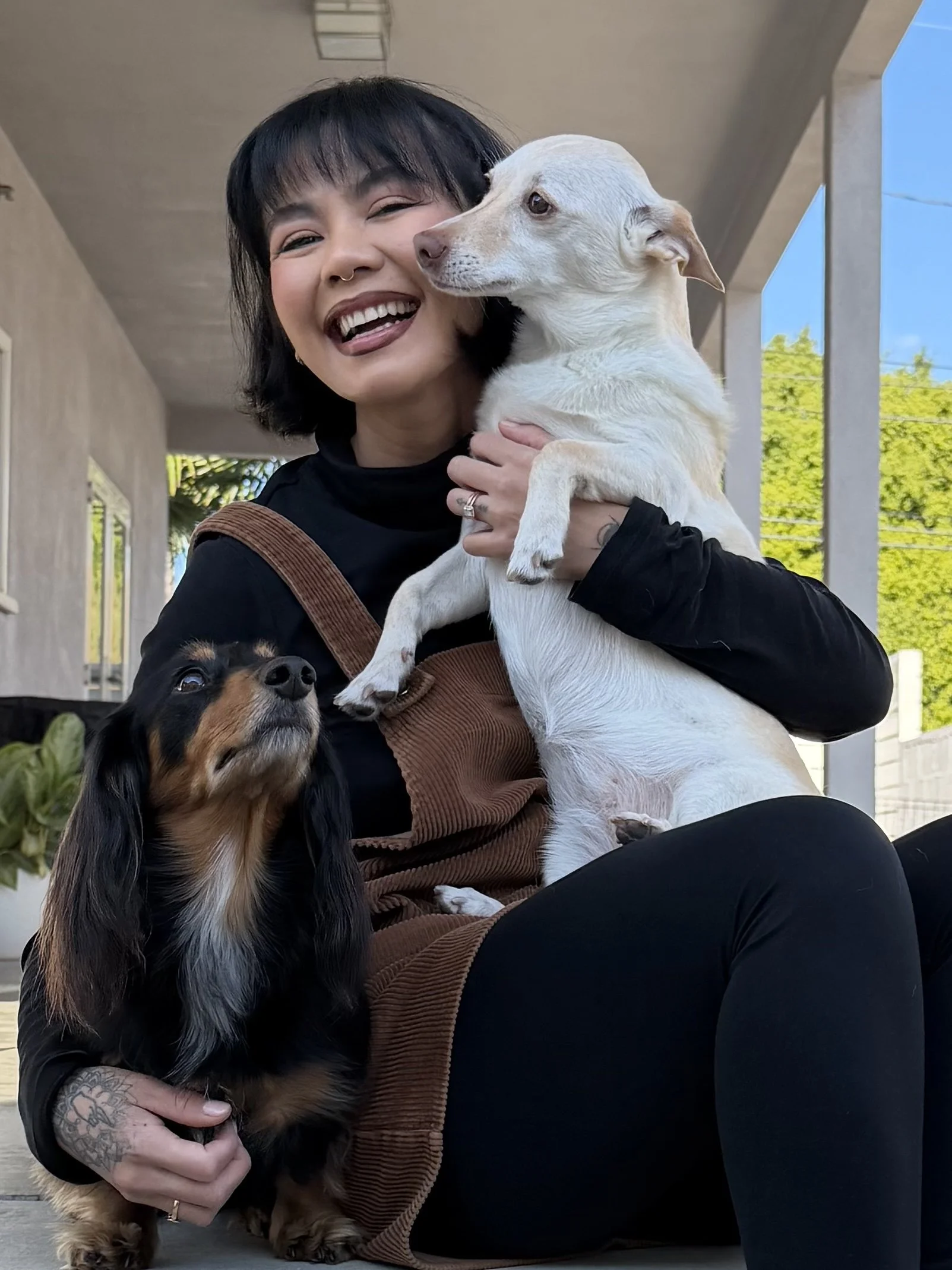 Rachelle smiling and holding a small tan and black dog under her arm, while a larger white dog with tan markings sits on her lap, looking to the side, outside a building with trees and a blue sky in the background.