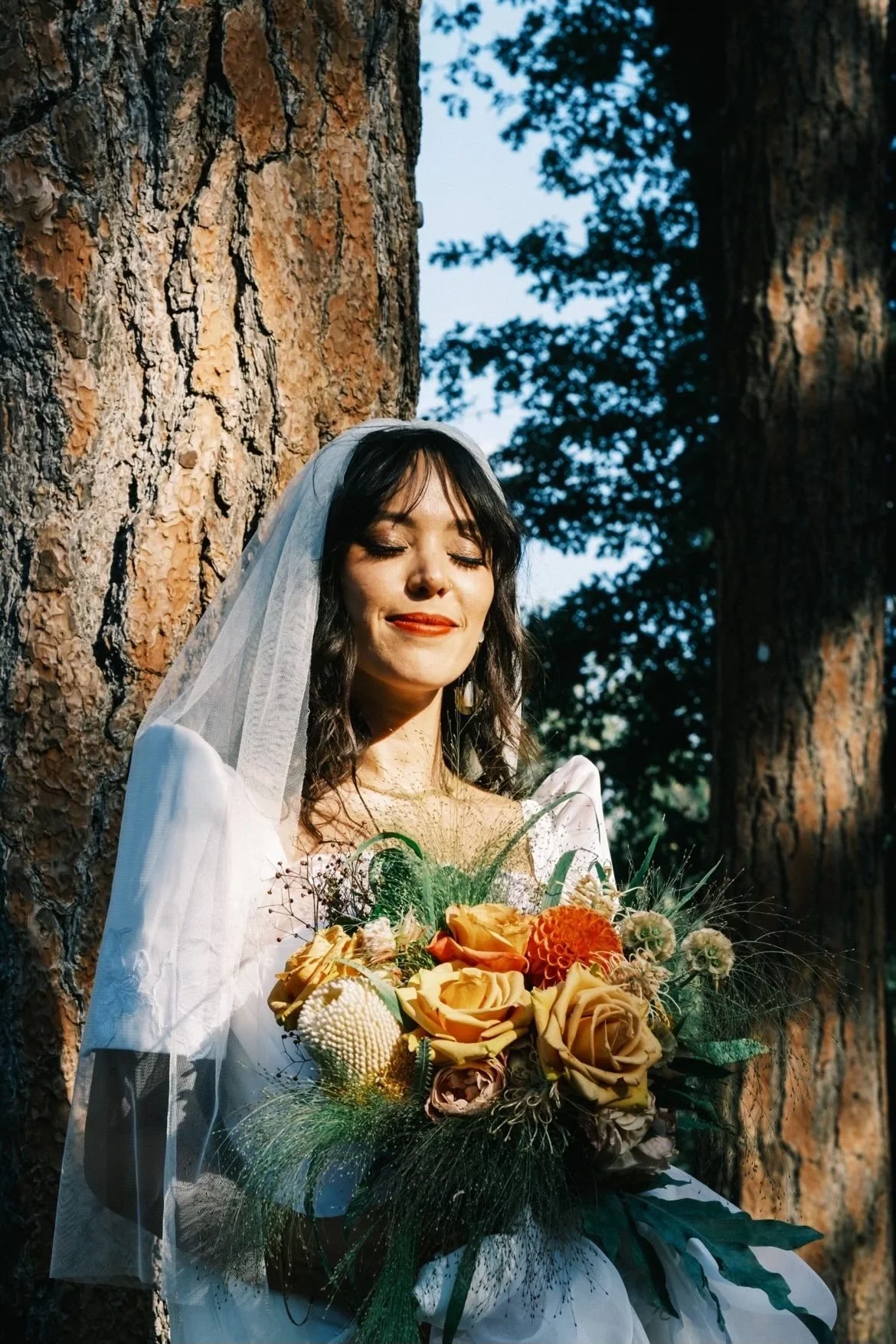 A woman in a white wedding dress and veil holding a bouquet of yellow and orange flowers, standing outdoors near large trees with sunlight filtering through.