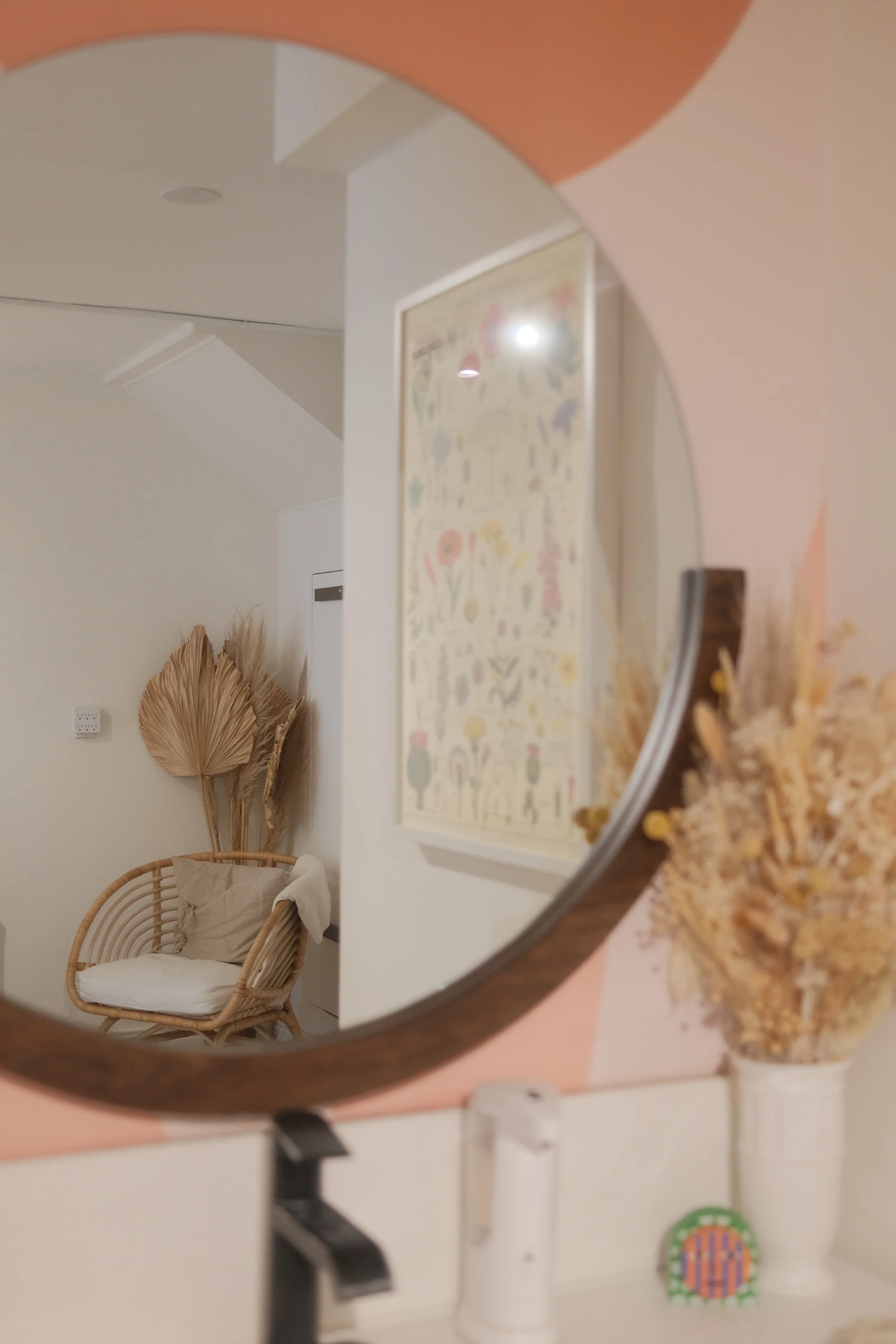 Reflection in a round mirror shows a white wicker chair with a cushion, framed artwork with floral patterns on the wall, dried decorative plants, and household items on a pink table, including a container with snacks and a small colorful object.