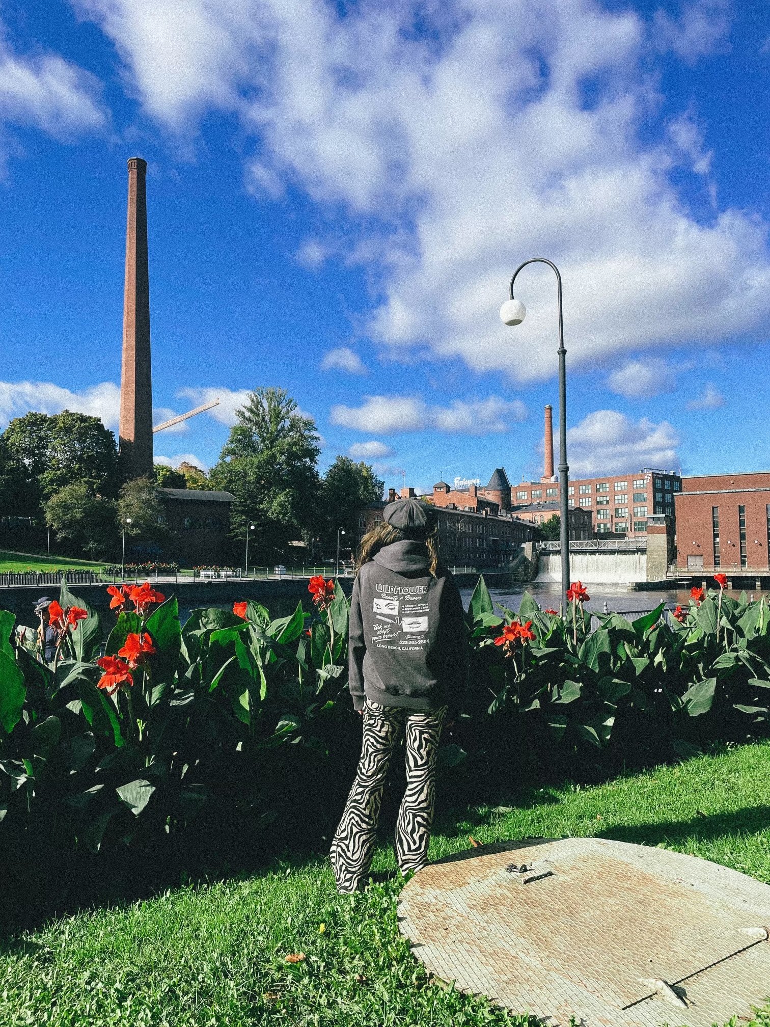 A person standing on grass near a river, facing away from the camera, wearing a hoodie with 'Wildflower' printed on the back and zebra-striped pants, surrounded by red flowers with a cityscape in the background under partly cloudy skies.