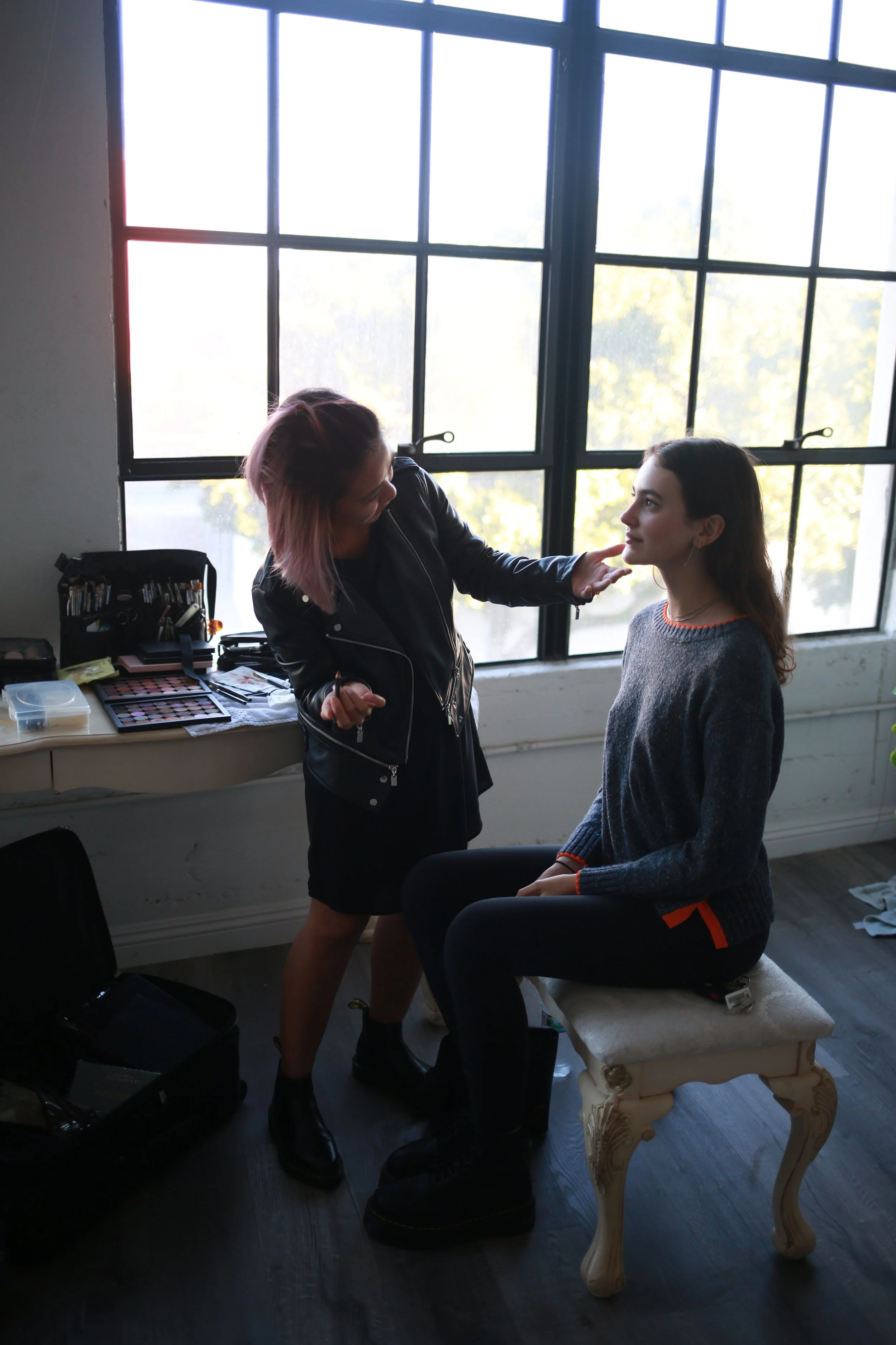 Makeup artist Rachelle applying makeup to a woman seated on a vintage-style stool near a large window.