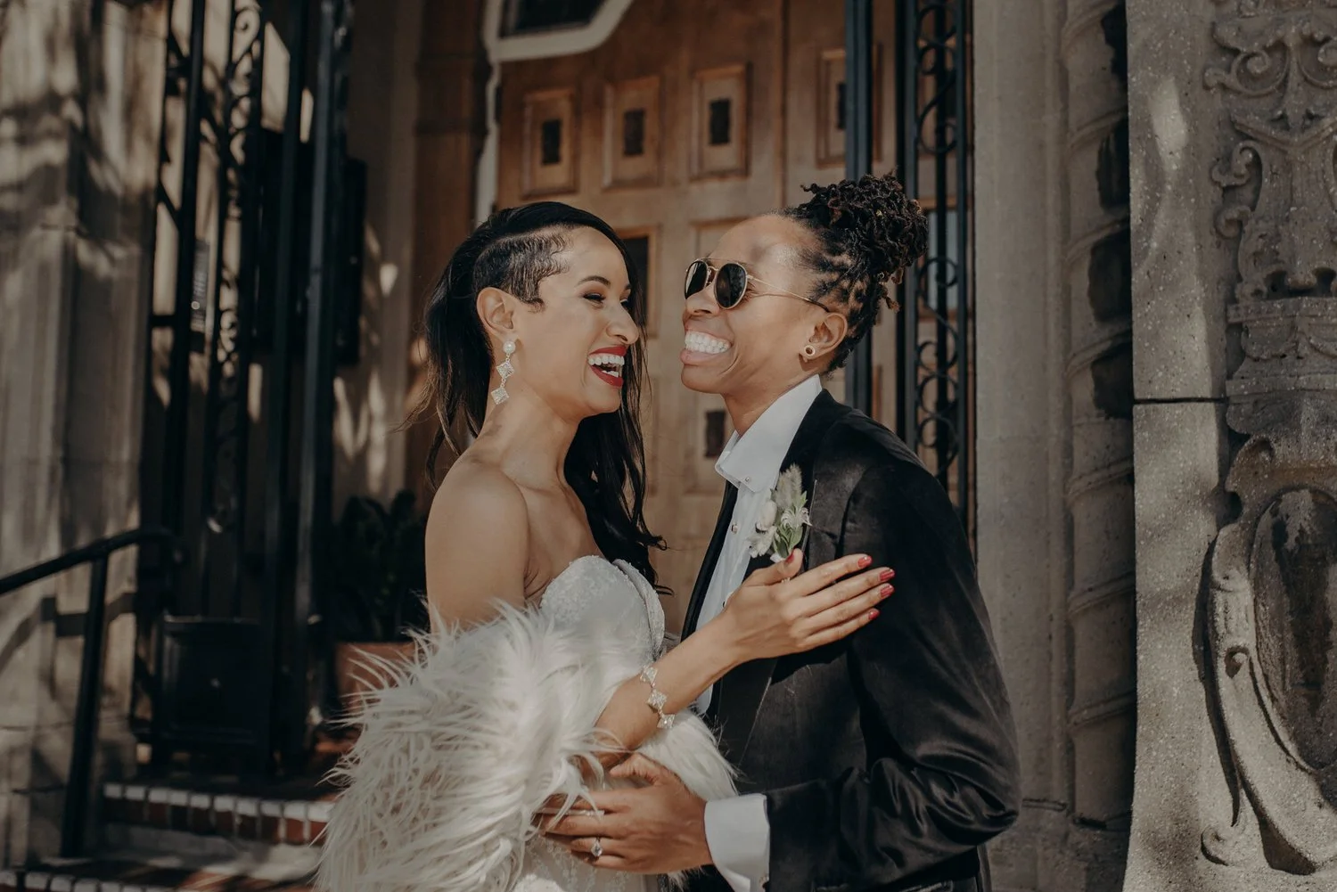 Two women dressed in wedding attire, smiling and embracing outside a building.