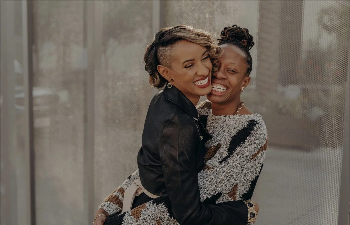 Two women hugging and smiling in front of a glass wall.