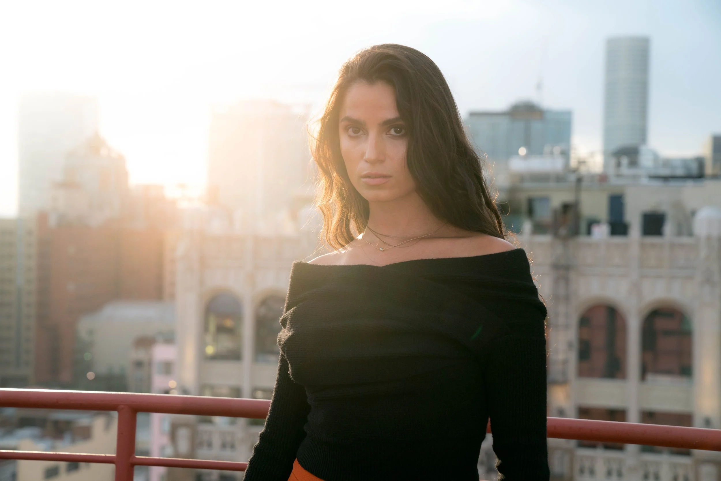 A woman with long dark hair and a serious expression stands on a balcony with a city skyline in the background during sunset.