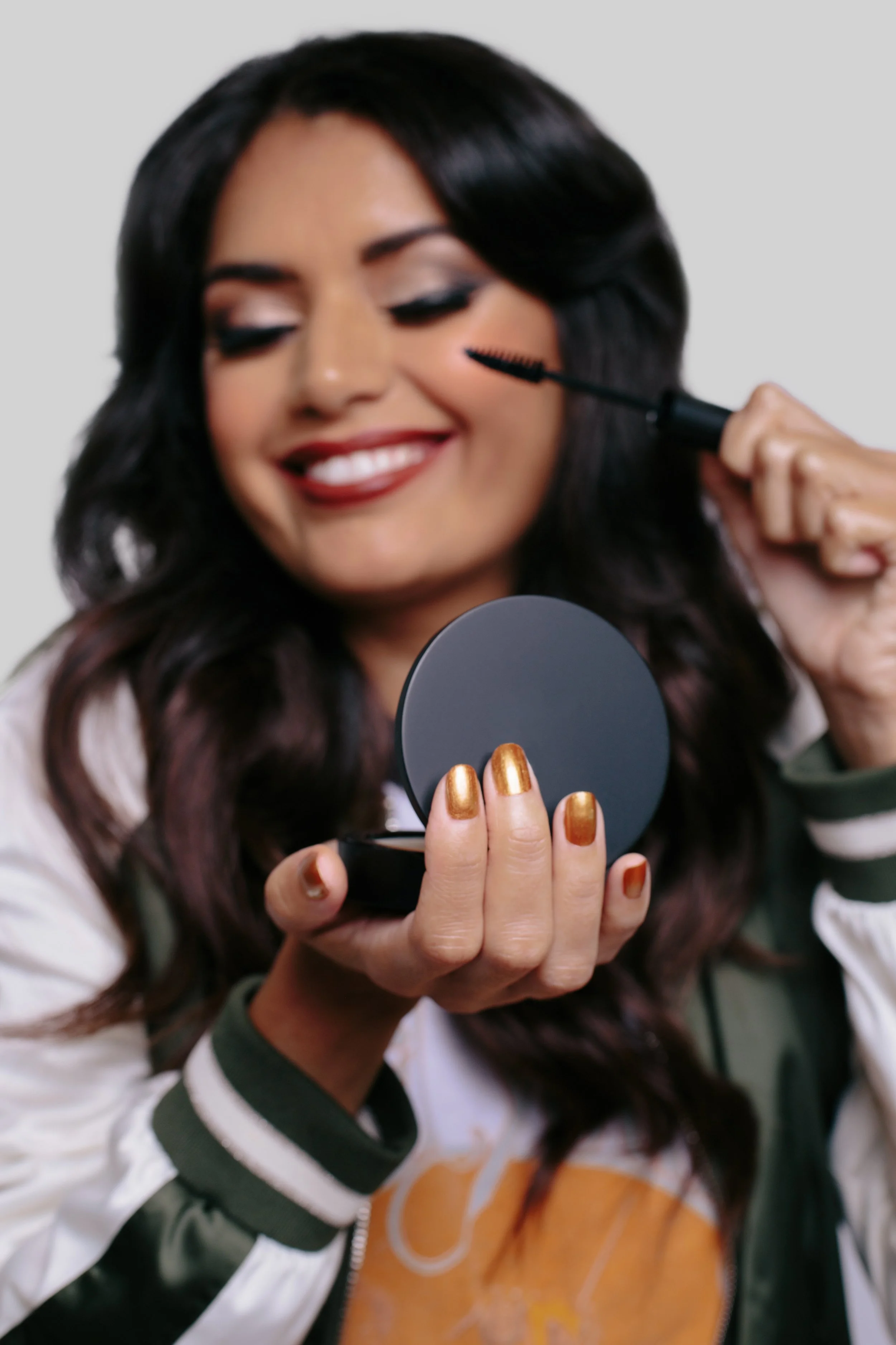 A woman with dark, wavy hair applying mascara while looking into a mirror, smiling, with a light gray background.