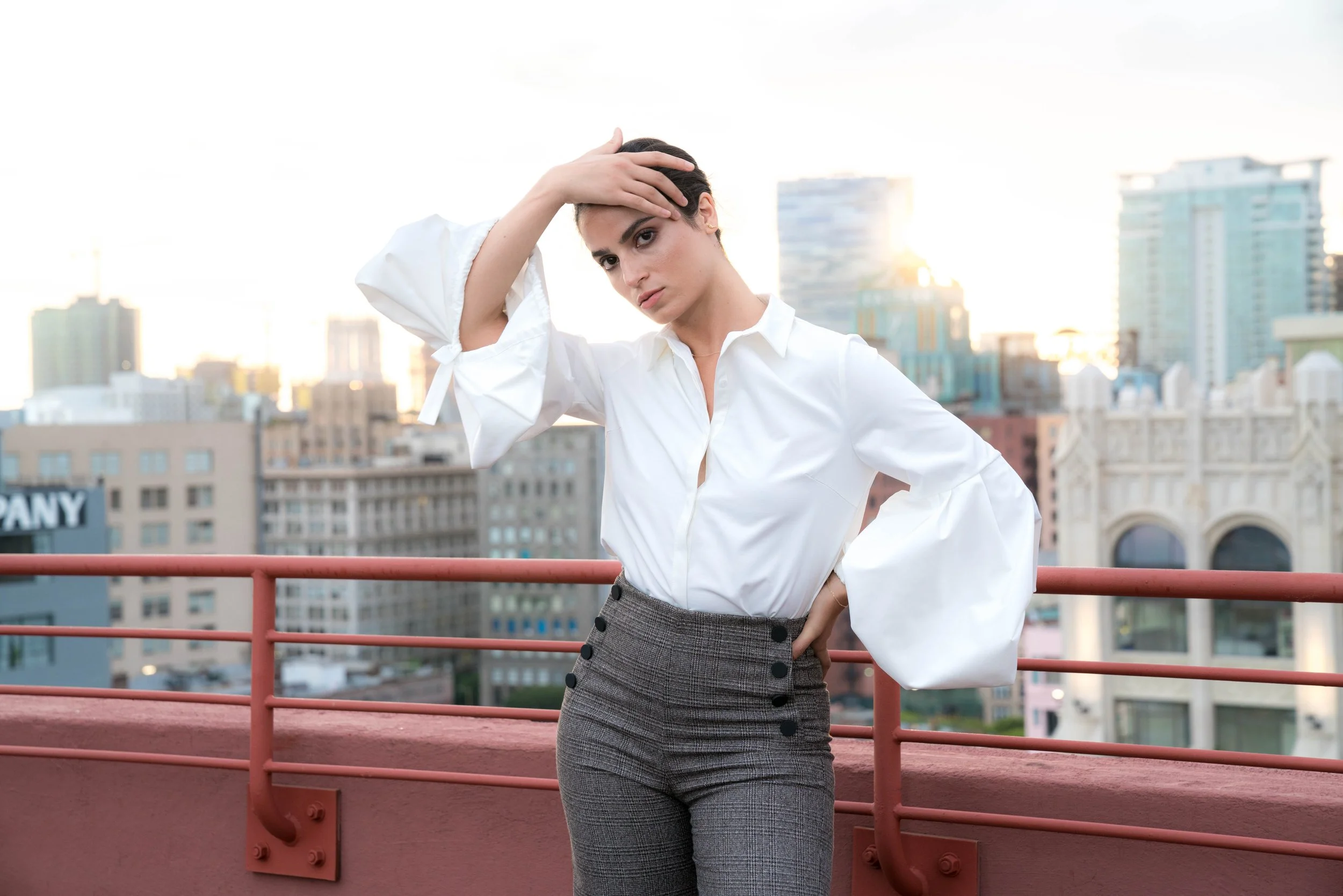 A woman in a white blouse and gray plaid skirt standing on a rooftop with city buildings in the background, holding her hair back with one hand.
