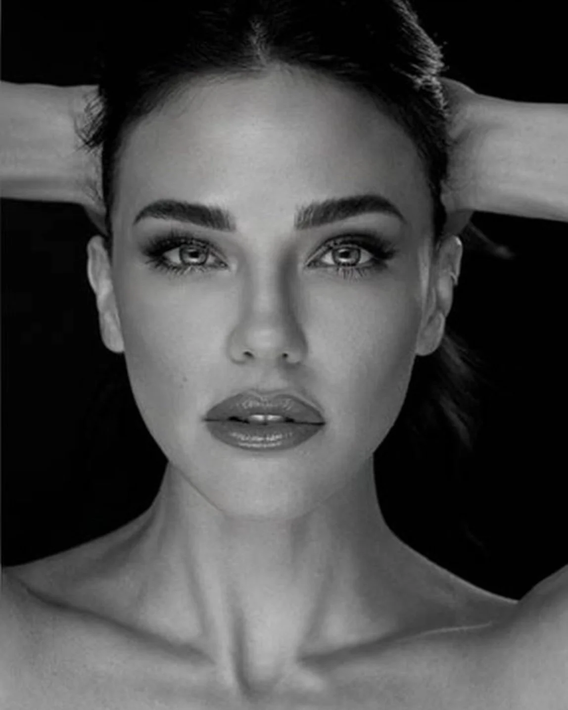Close-up black and white photograph of a woman with dark hair, striking eyes, full lips, and a confident expression, with her hands behind her head.