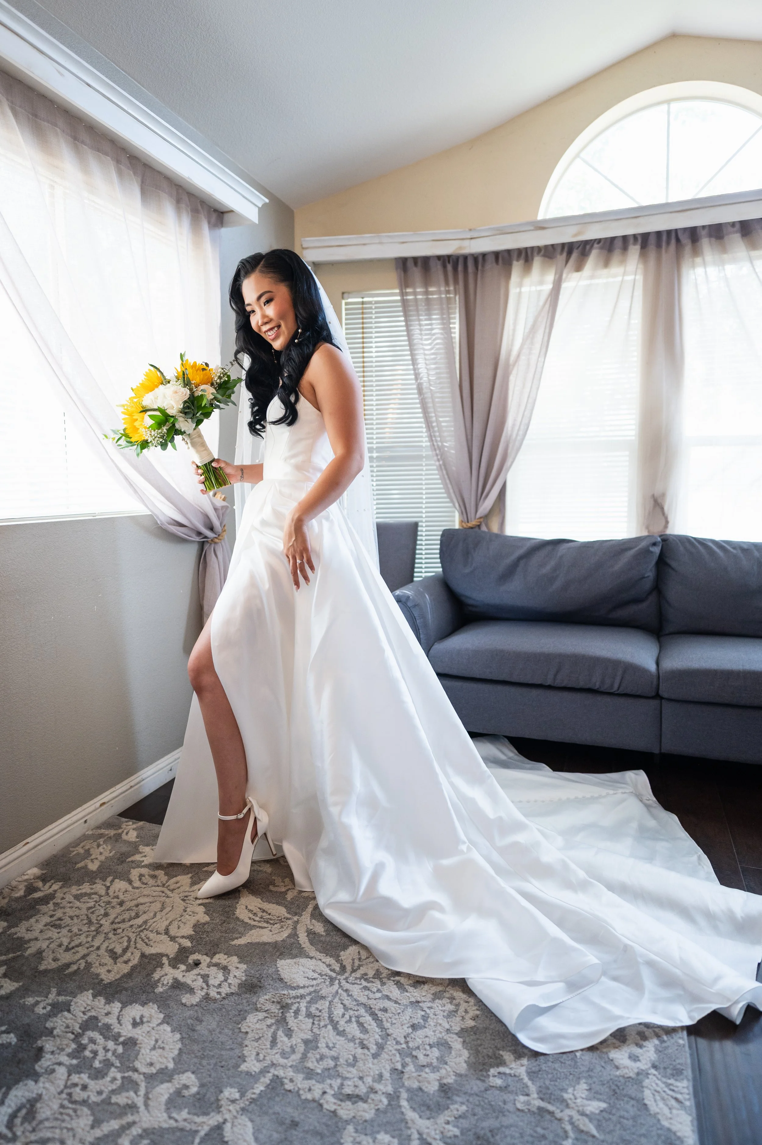 Brunette woman in a wedding dress holding a bouquet of sunflowers and white flowers smiling in a room with large windows and gray curtains.
