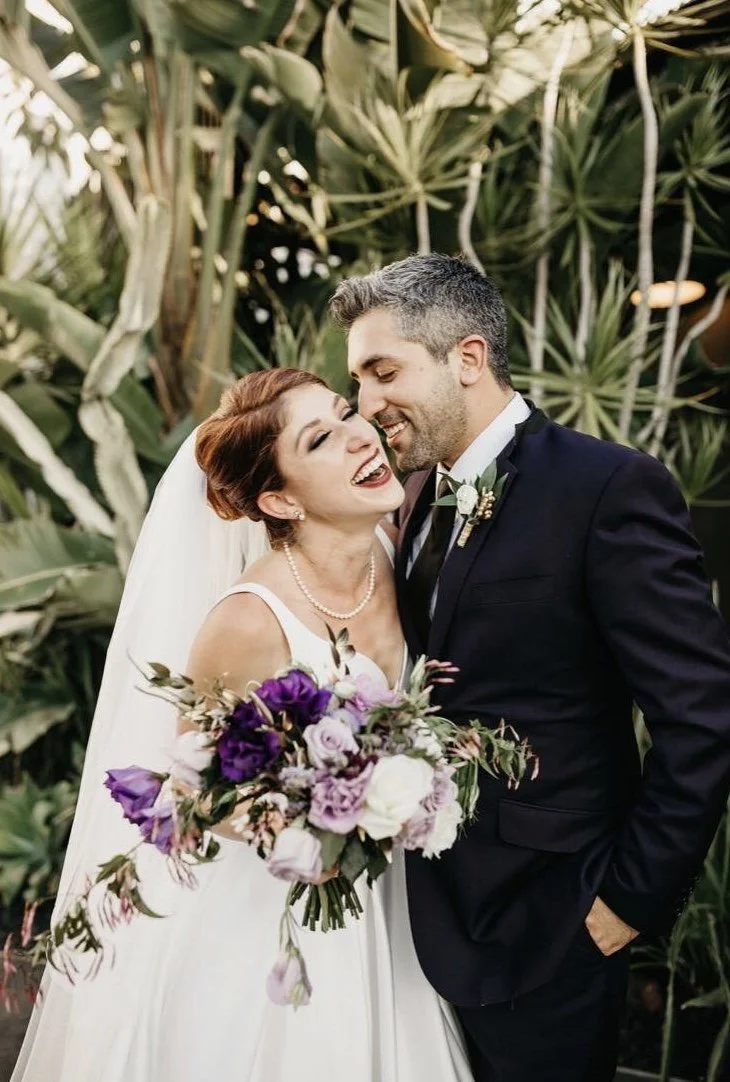 A bride and groom celebrating their wedding outdoors with lush greenery in the background. The bride is smiling and holding a bouquet of purple and white flowers. The groom is dressed in a dark suit with a boutonniere, leaning towards the bride.
