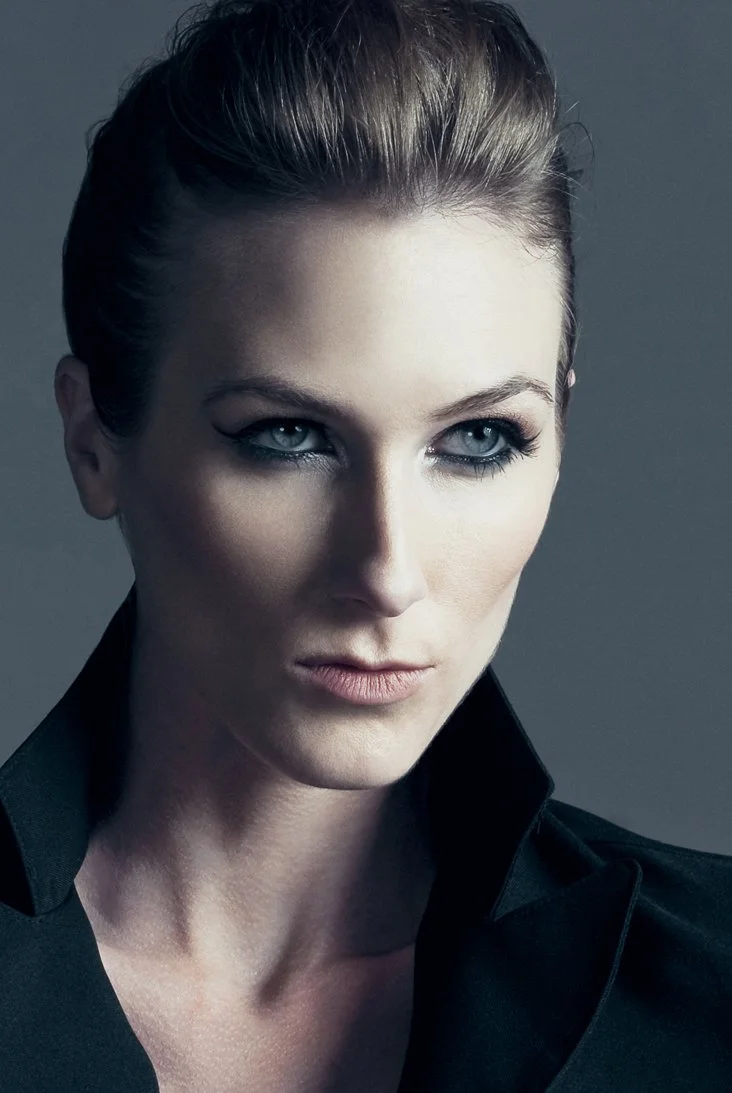 Close-up of a woman with blonde hair styled back, wearing a black jacket with a raised collar, and looking off to the side with a serious expression.