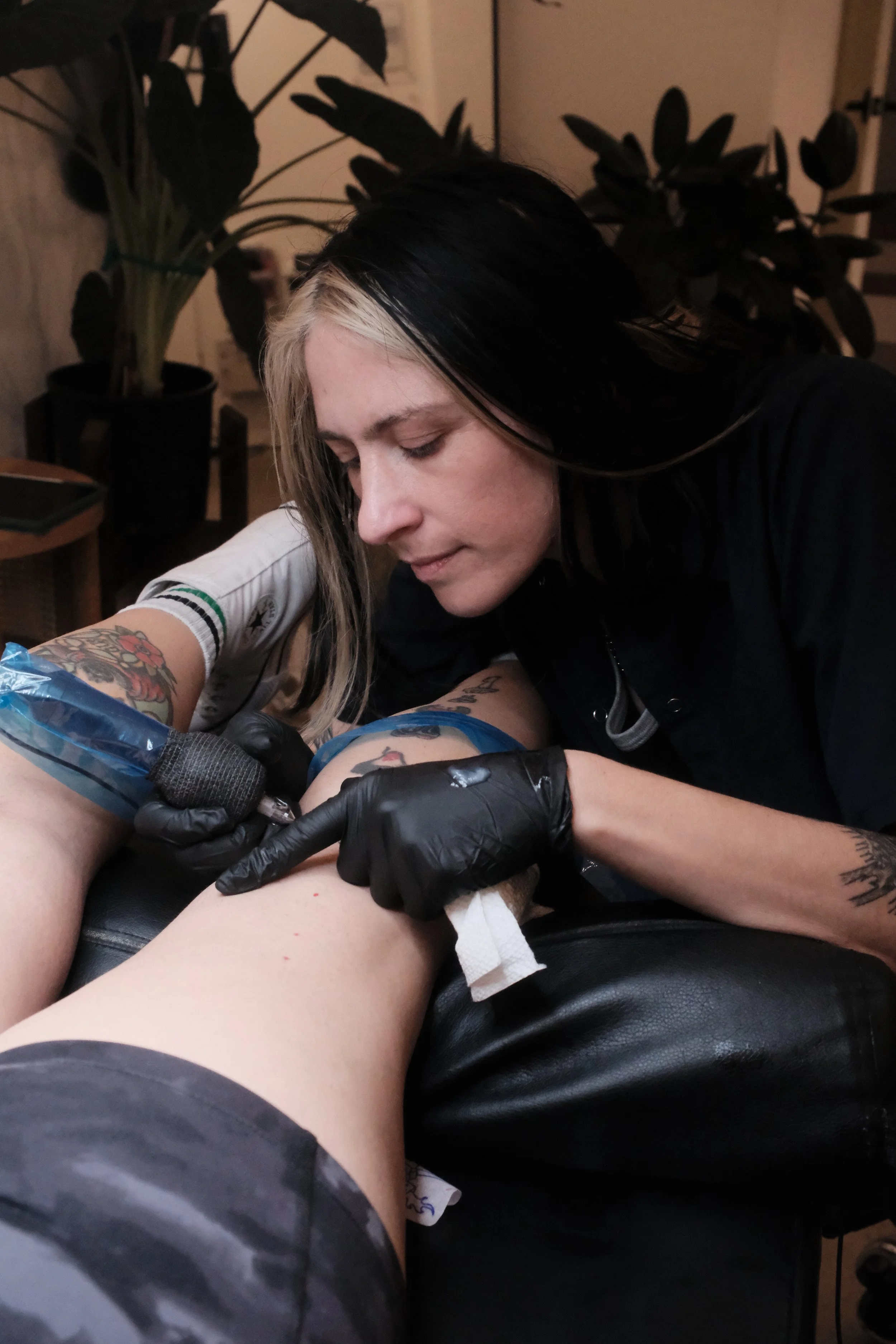 Garnet is tattooing another person's leg. The focus is on the tattooing process, with a tattoo machine in hand. The person receiving the tattoo is lying on a black surface