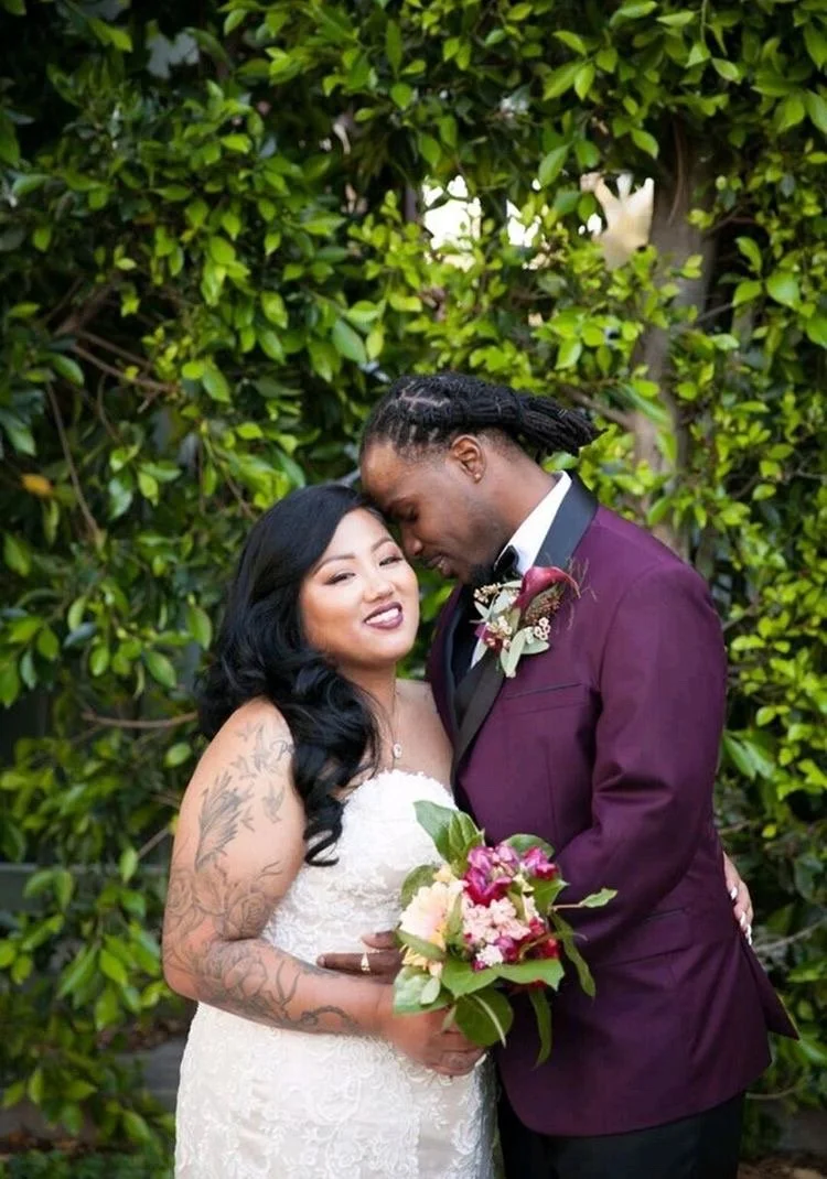 A newlywed couple, the bride and groom, embrace outdoors in front of green foliage. The bride, with long black hair and tattoos on her arm, holds a bouquet of flowers. The groom, dressed in a purple tuxedo, gently rests his forehead against the bride
