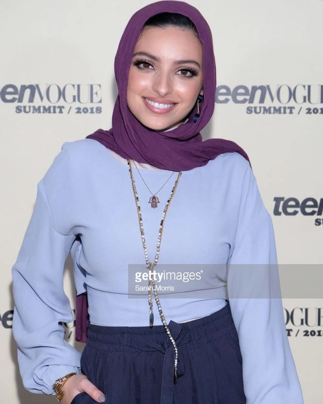 A young woman wearing a purple headscarf, white top, and dark pants smiling at a fashion event with a Teen Vogue backdrop.