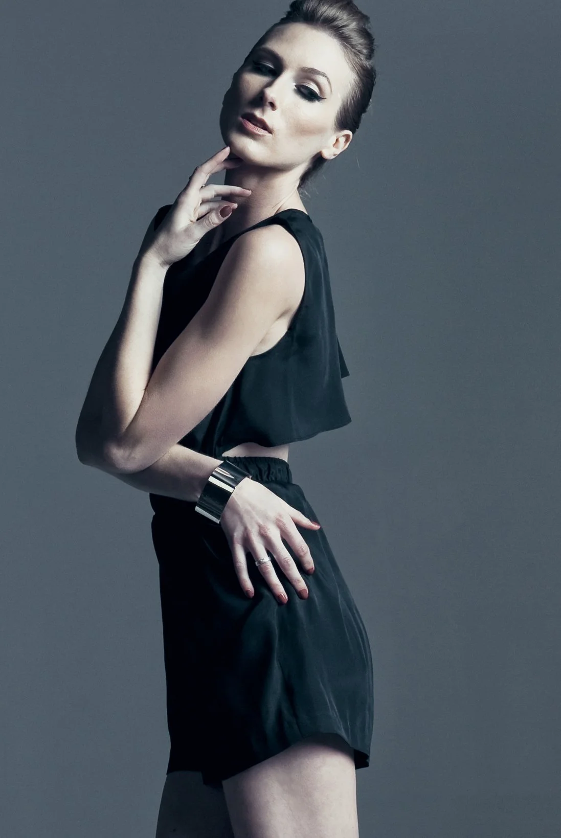 A woman wearing a black sleeveless top and shorts, accessorized with a silver bracelet and rings, poses with her hand near her face against a gray background.