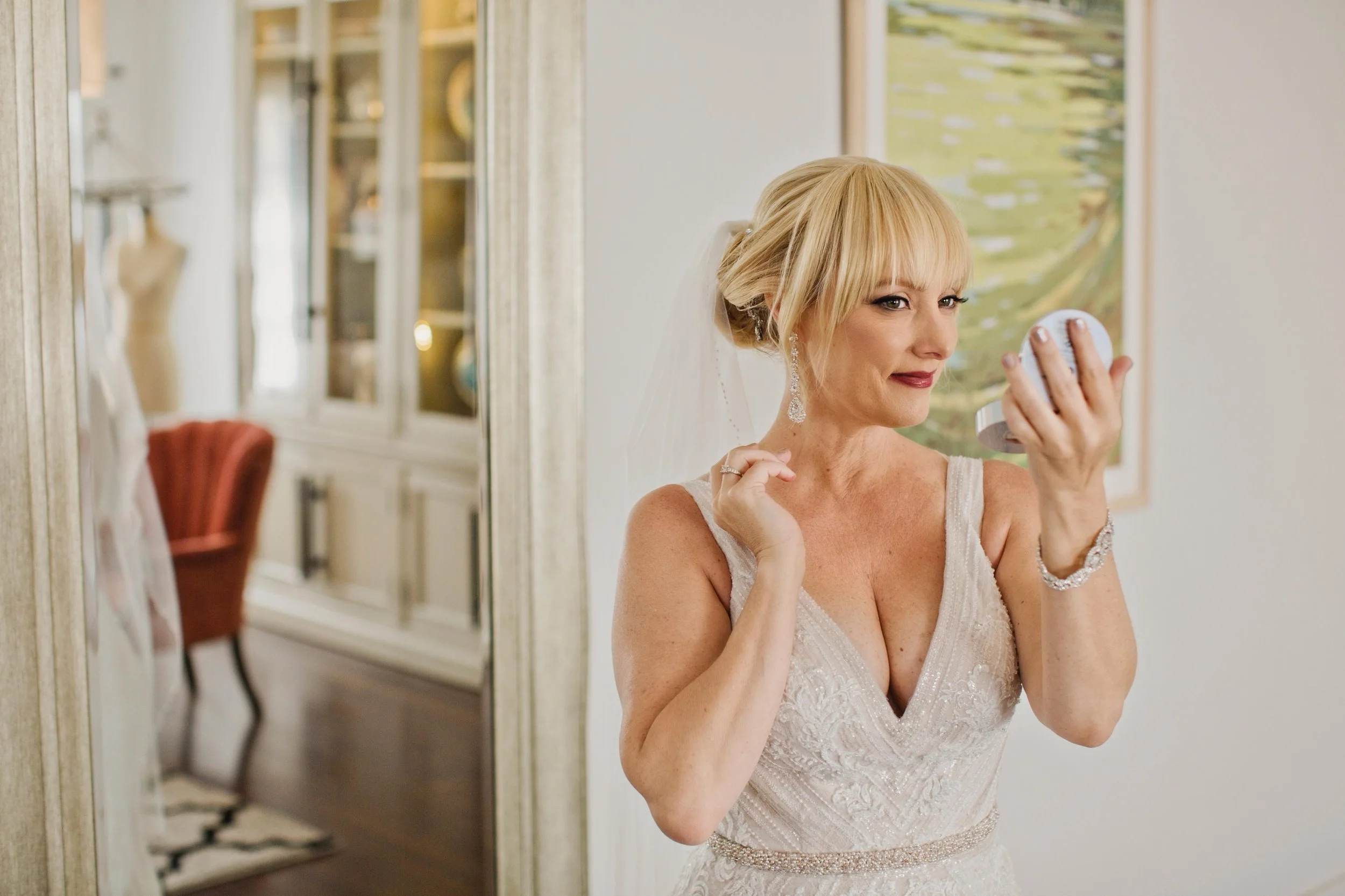 A woman with blonde hair in an updo wearing a white lace wedding dress, earrings, and jewelry, applying perfume in front of a mirror in a well-lit room.