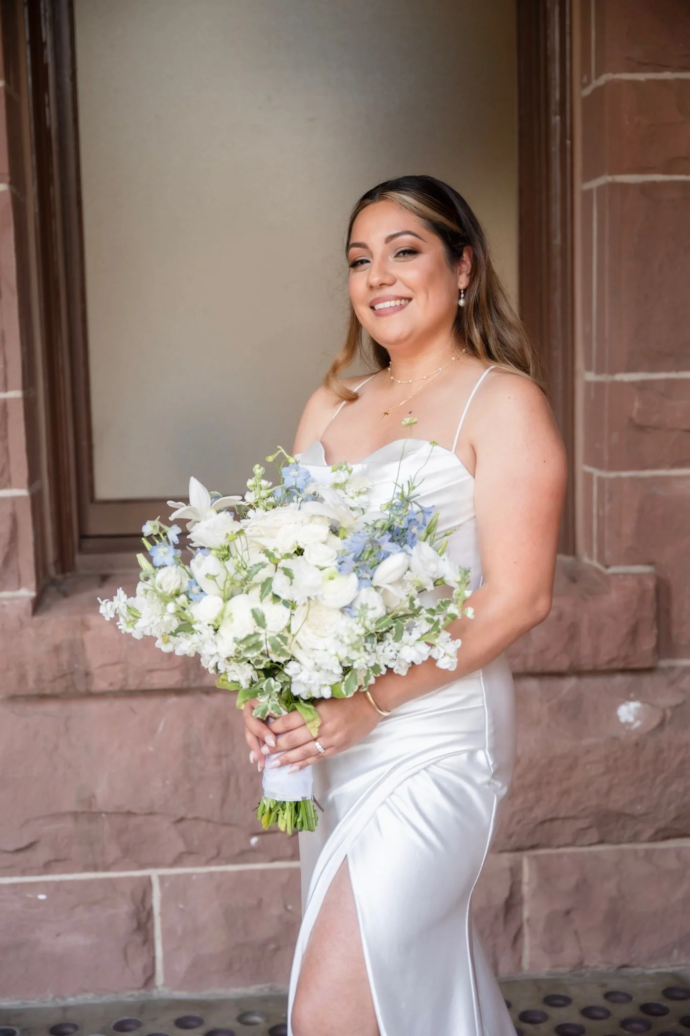 A woman in a white dress with spaghetti straps holding a large bouquet of white and light blue flowers, standing in front of a brown brick wall. She is smiling and winking at the camera.