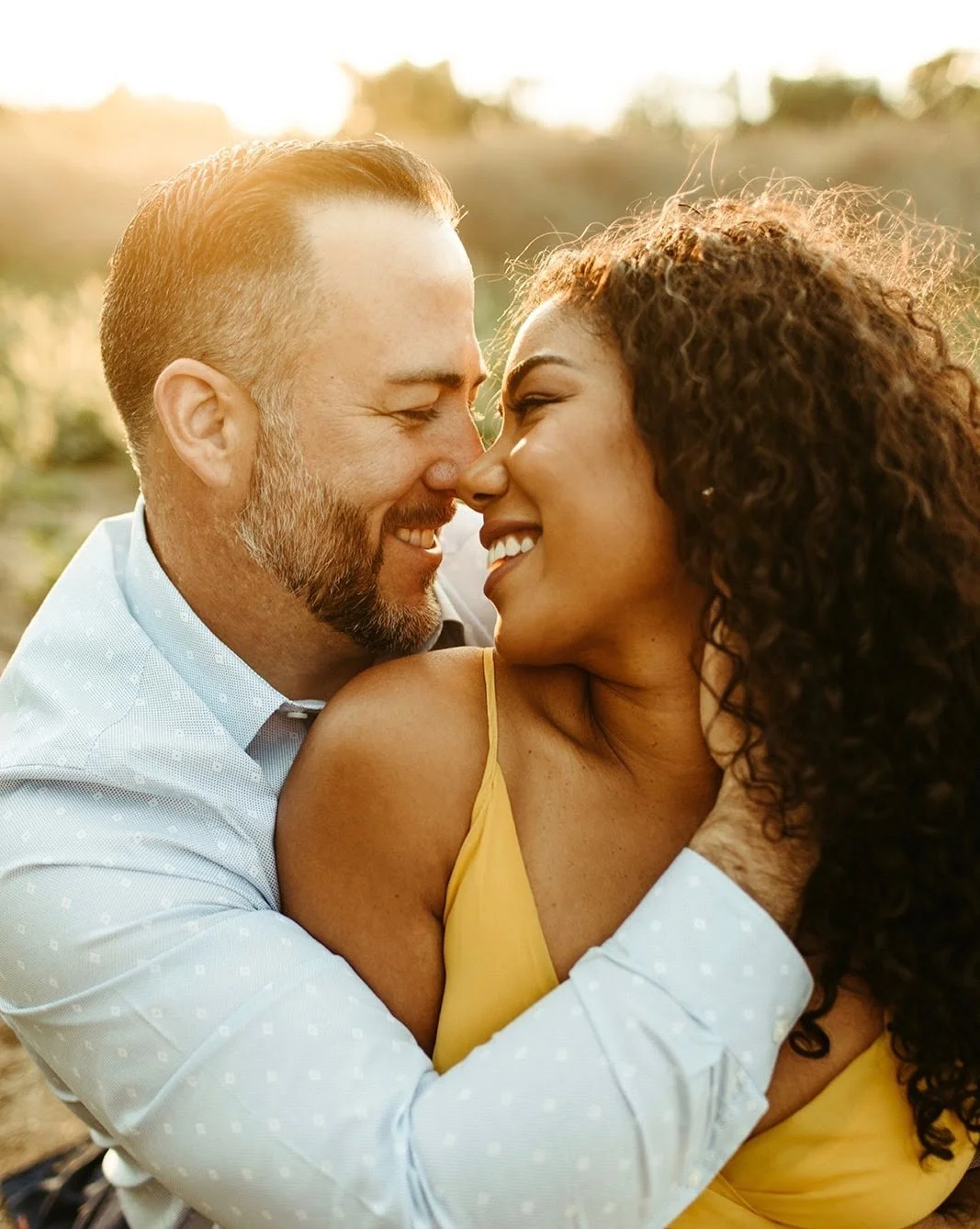A loving couple smiling and touching noses outdoors during sunset, with blurred greenery in the background.