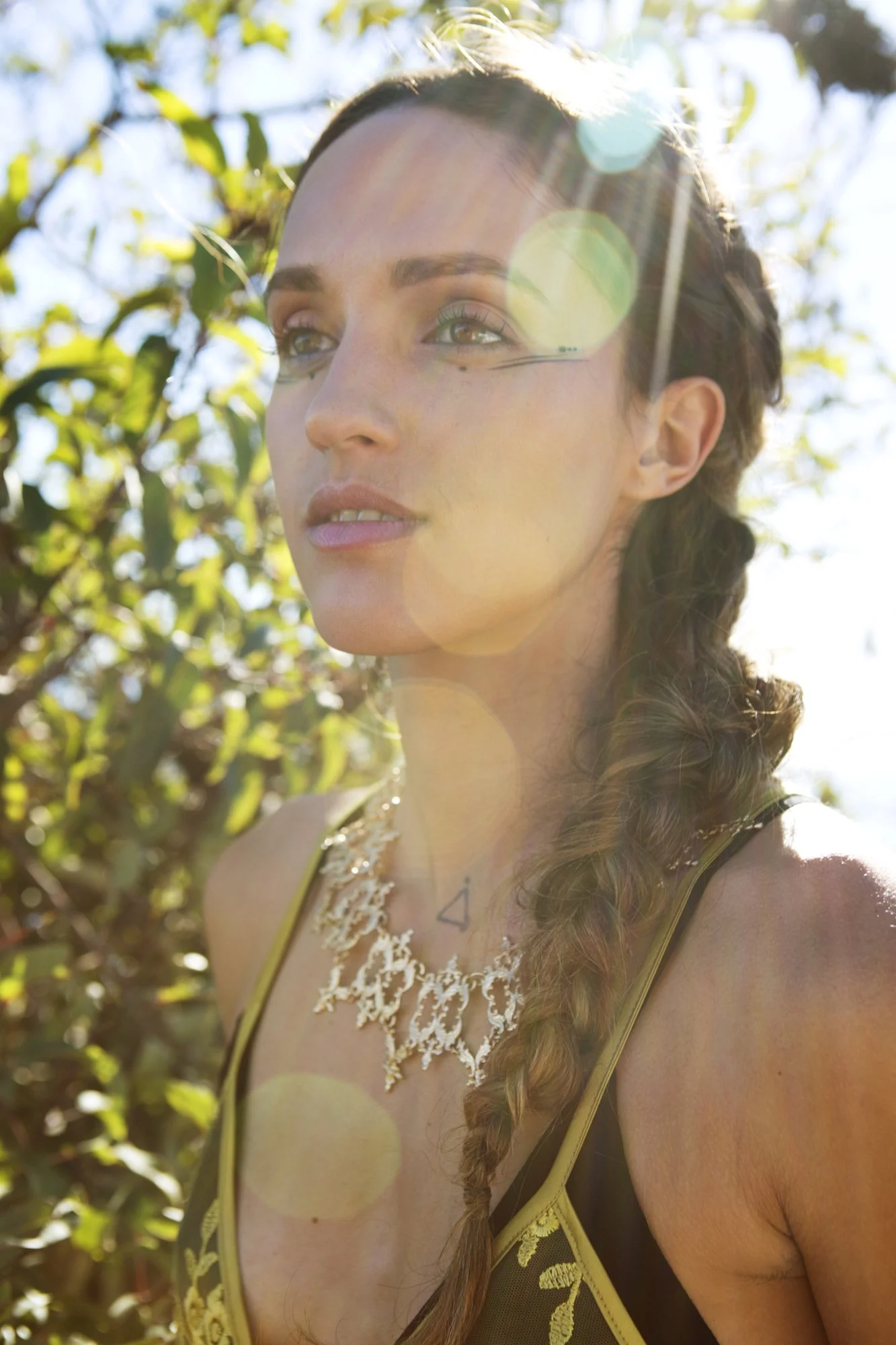A woman with brown hair and green eyes in a braid, wearing a black top with yellow embroidery, a lace necklace, and black eyeliner, standing outdoors in sunlight with a background of trees and leaves.