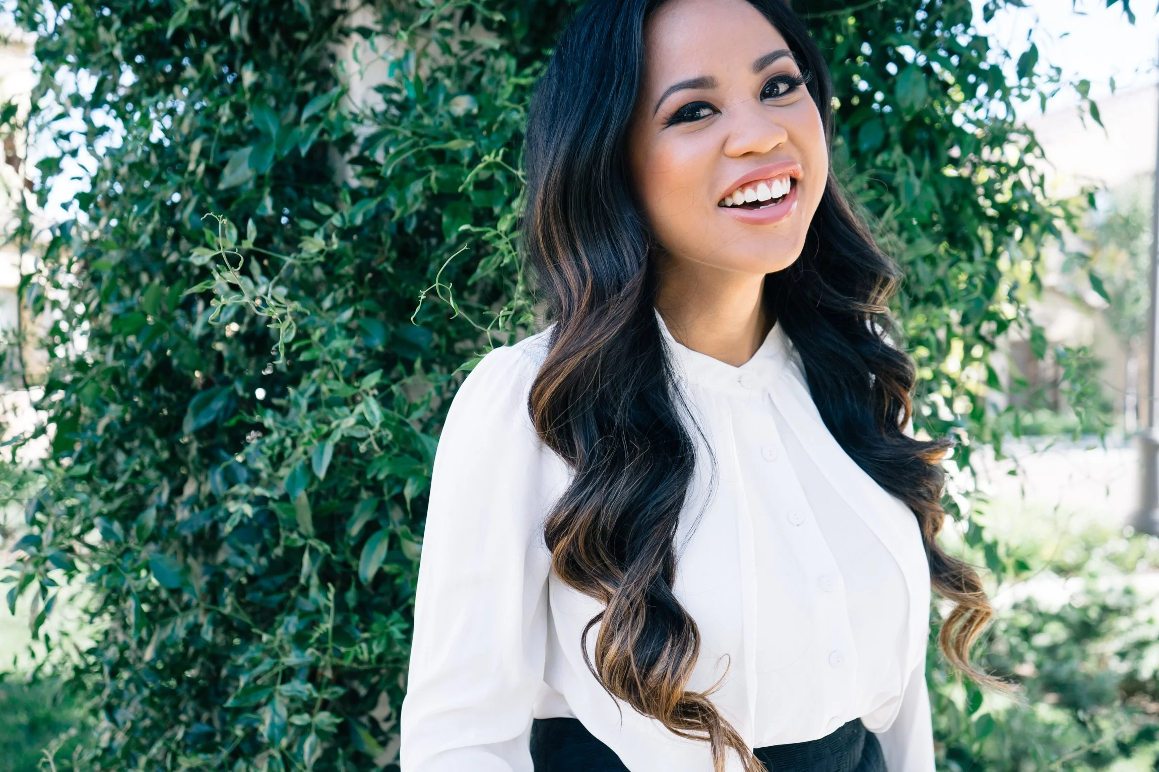 A woman with long dark hair and a white blouse smiling outdoors in front of greenery.