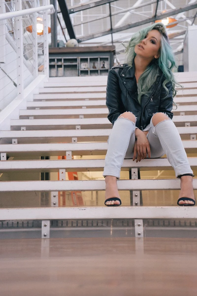Woman with teal hair sitting on stairway in industrial-style space, wearing a black leather jacket, ripped white jeans, and black heels.