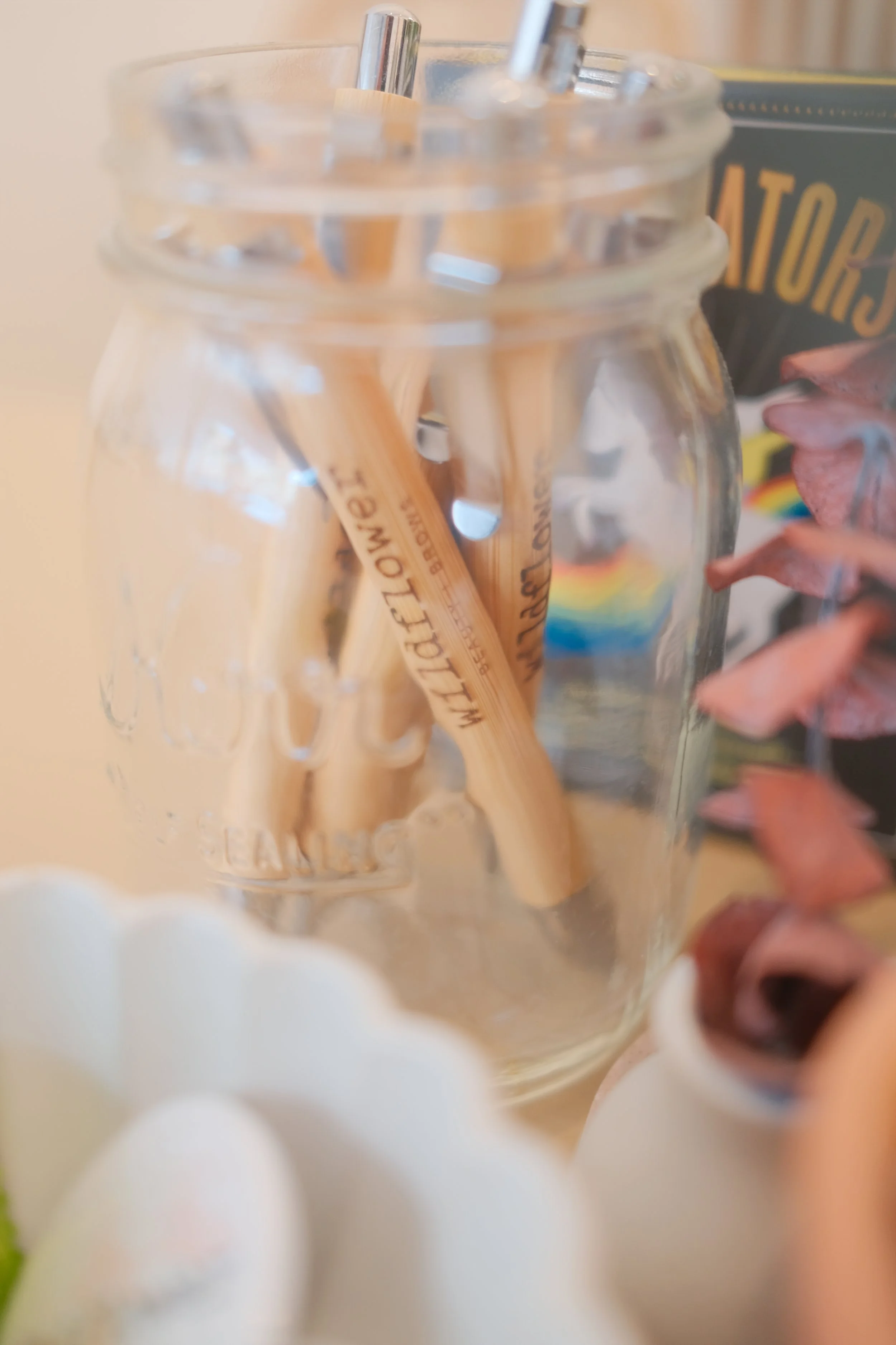 Close-up of a glass jar filled with wooden clothespins, with a book titled 'The Artist' partially visible in the background, and pink dried flowers on the right side.