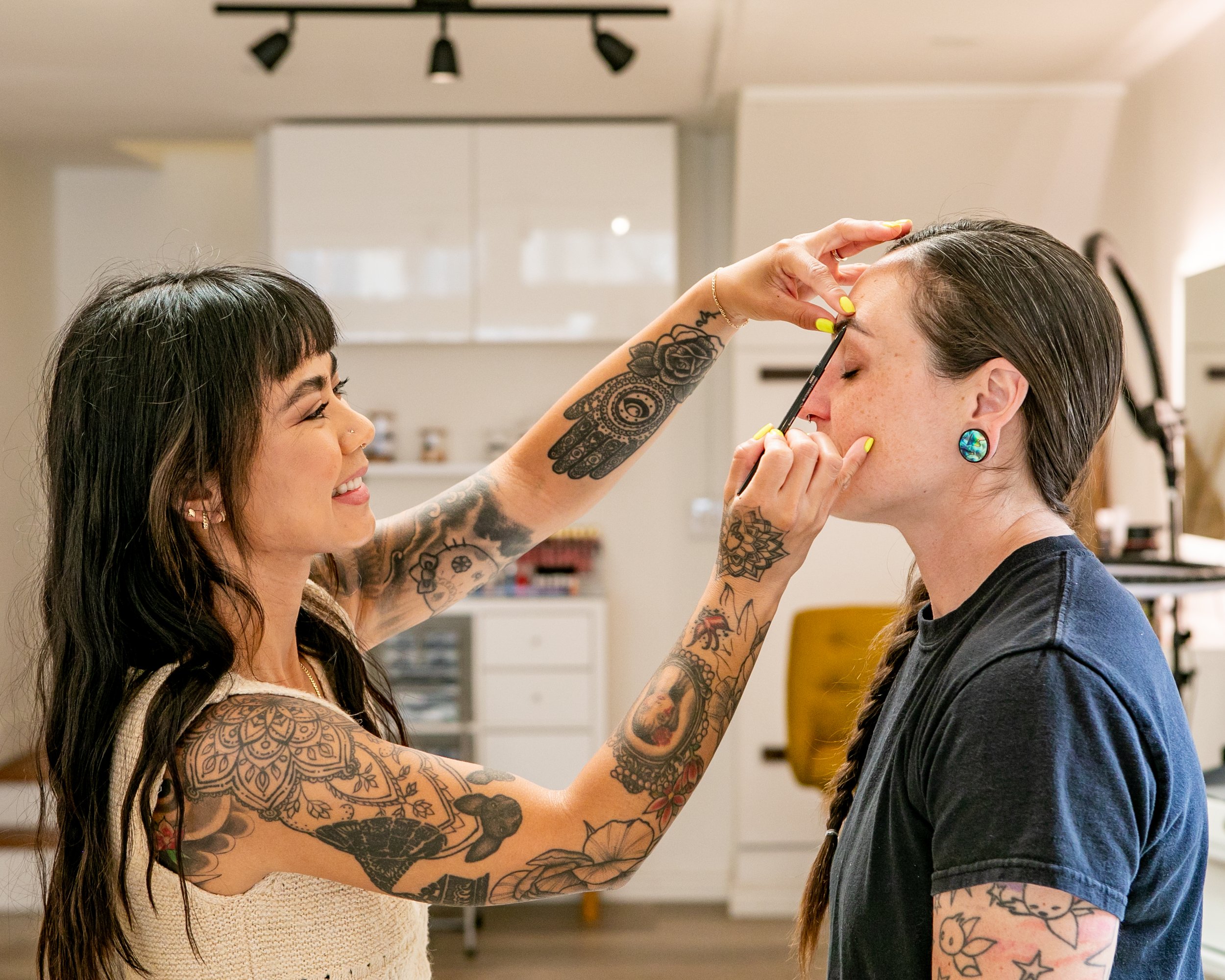 Rachelle with tattoos and dark hair applies makeup to a woman with dark hair and colorful earrings, in a room with white walls and makeup supplies.