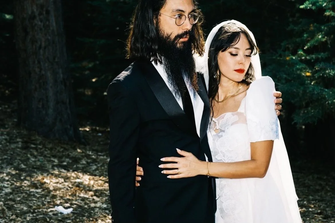 A couple dressed in wedding attire embracing outdoors in a wooded area, with the woman wearing a white dress and the man in a black suit with glasses and long hair.