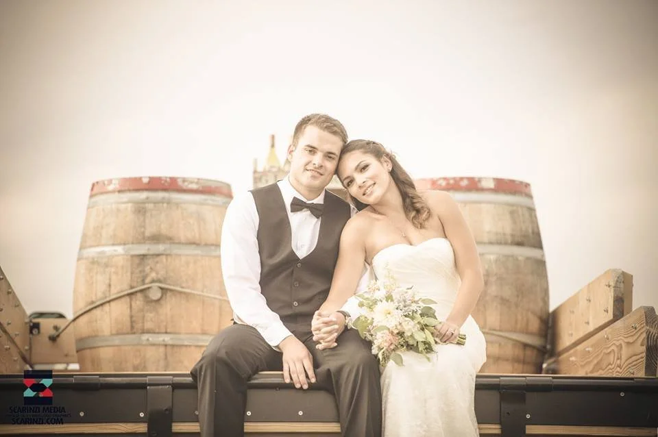 A newlywed couple sitting on a wooden trailer, holding hands with large wine barrels in the background, and the bride holding a bouquet.