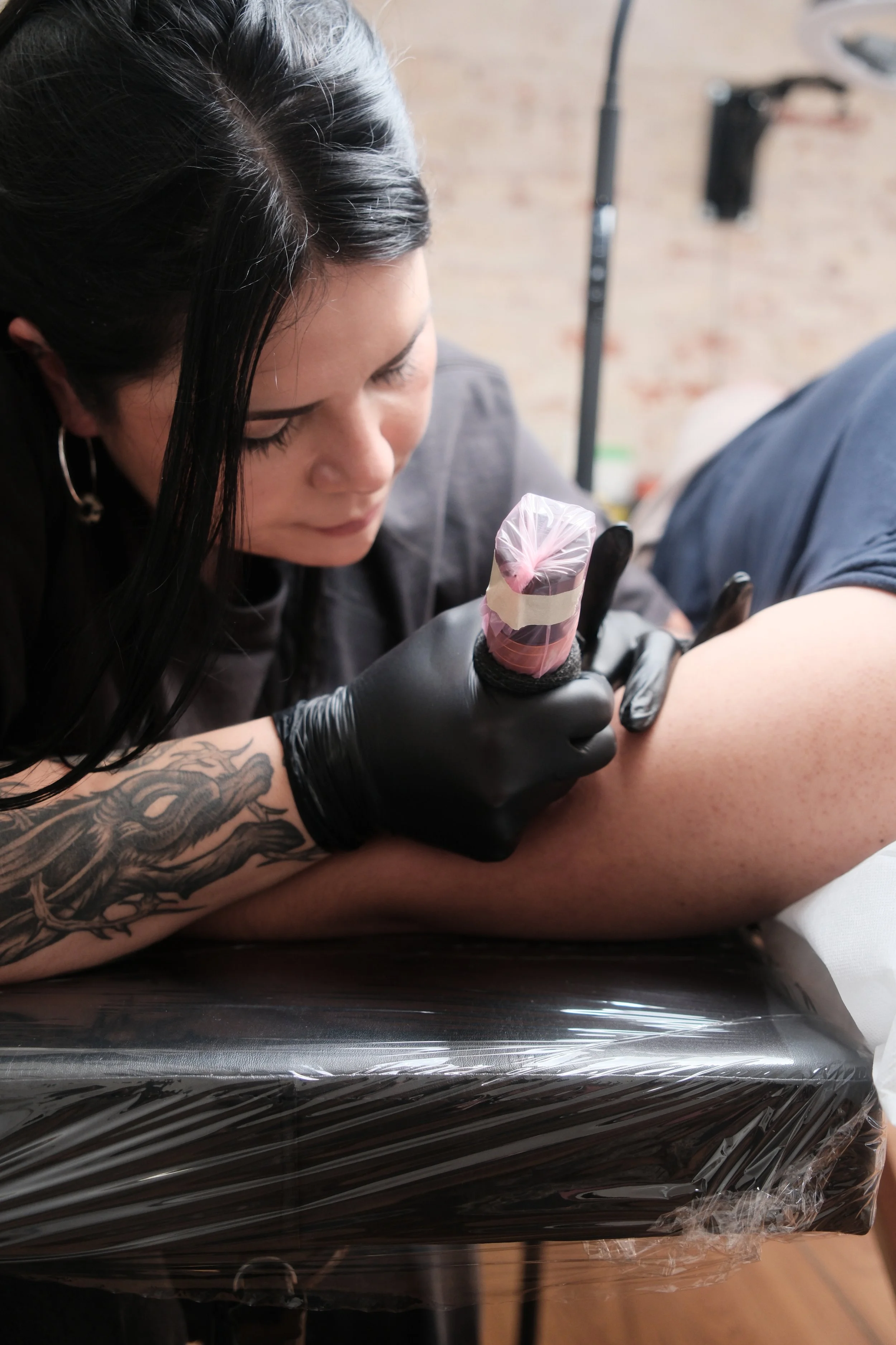 Samantha wearing black gloves, is tattooing a person's arm. The person receiving the tattoo has light skin, and their arm is resting on a black surface covered with plastic. The background shows a brick wall and some tattooing equipment.