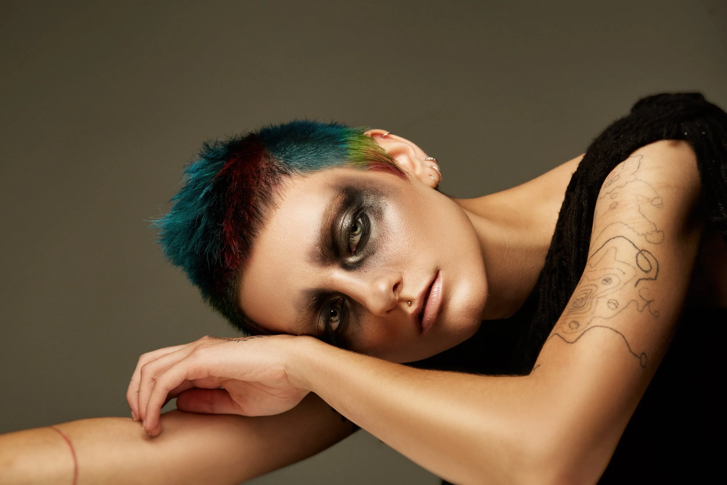 A person with short, multicolored hair lying on their side with their head resting on their arm, looking into the camera. They have dark makeup around their eyes, a septum piercing, and tattoos on their arm. The background is plain and neutral.