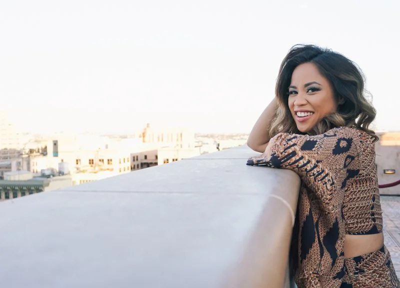 Woman with dark wavy hair and a brown patterned top smiling and leaning on a rooftop railing with city buildings in the background.