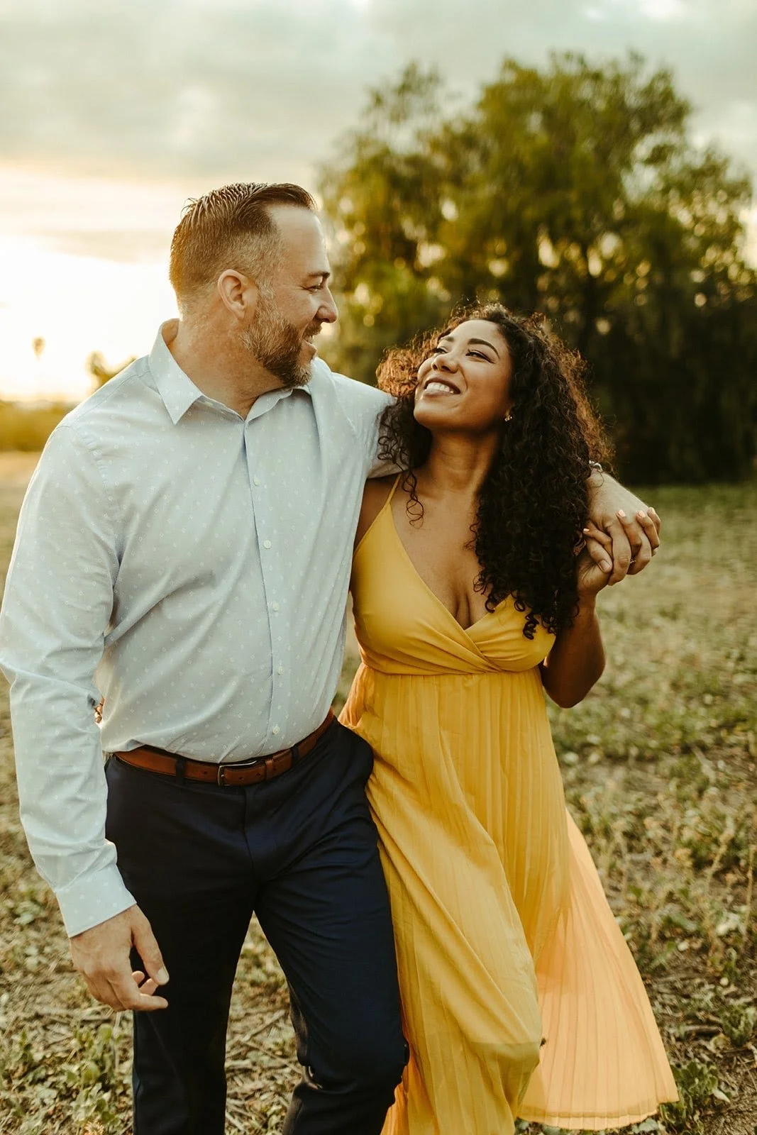 A man and woman walking together outdoors at sunset. The man is wearing a light blue shirt and dark pants, and the woman is wearing a yellow dress. They are smiling and looking at each other, with trees in the background.