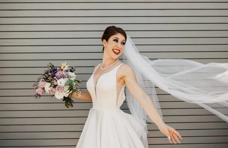 A woman in a wedding dress holding a bouquet of flowers with a big smile, standing in front of a gray, horizontal panel background.
