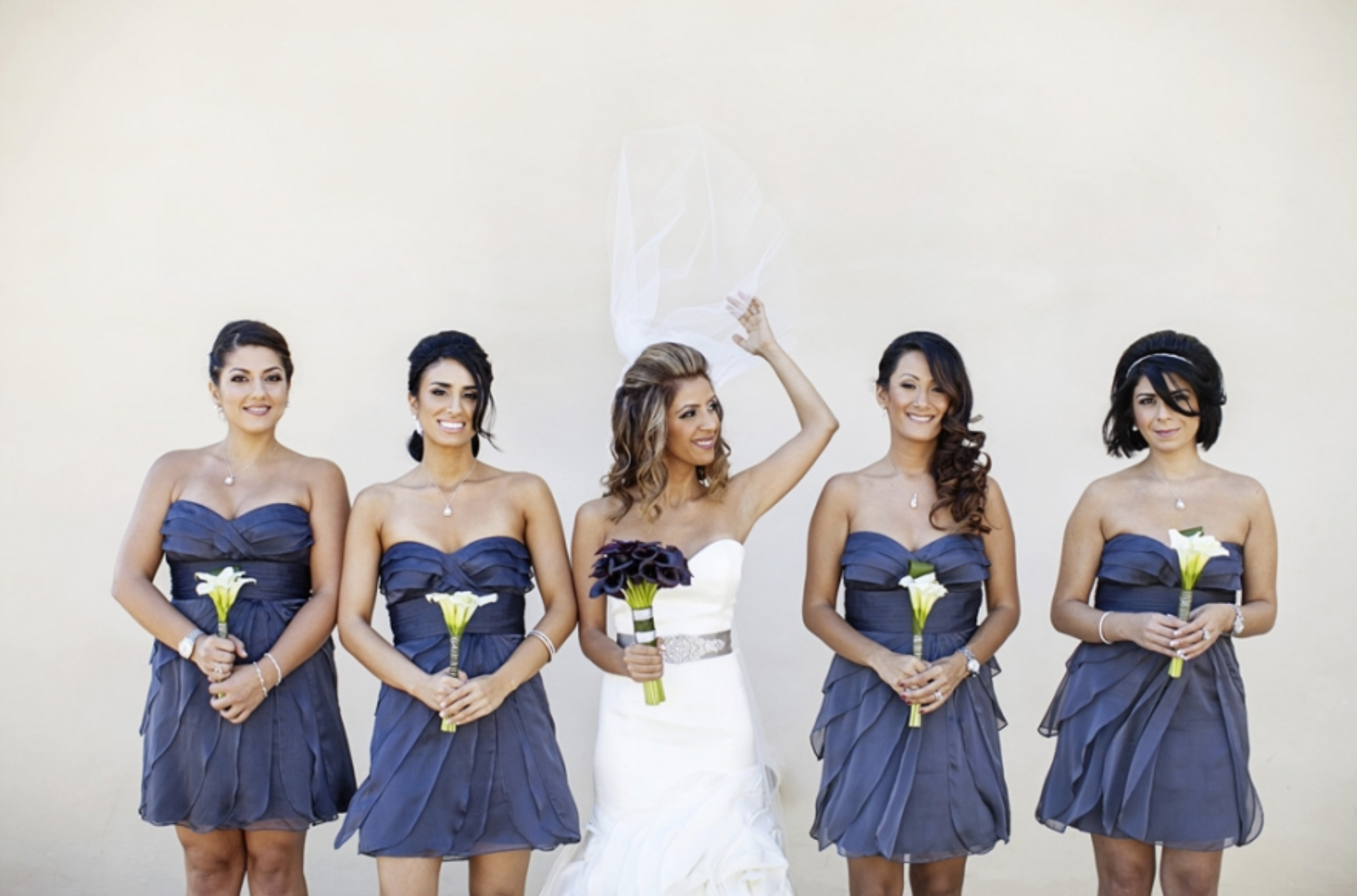 A bride and four bridesmaids standing in front of a plain light-colored wall. The bride is in the middle, wearing a white wedding dress, holding a bouquet of dark purple calla lilies, and tossing a piece of white tissue paper in the air. The bridesma