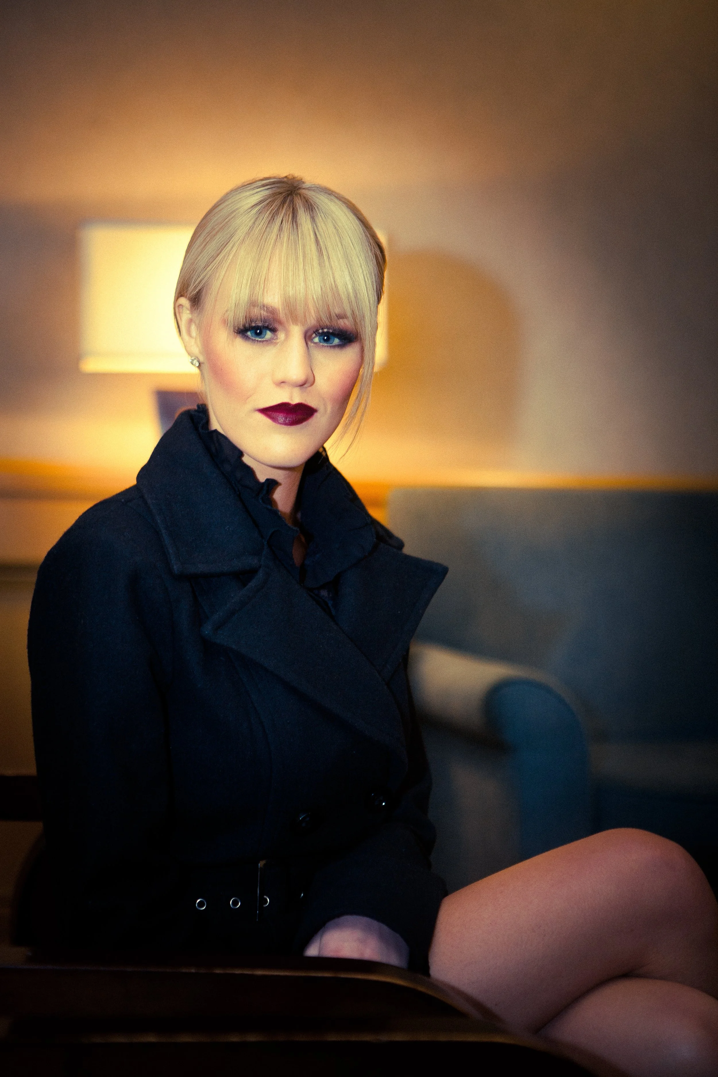 A blonde woman with blue eyes wearing dark lipstick and a black coat sitting indoors near a lamp with a beige lampshade.