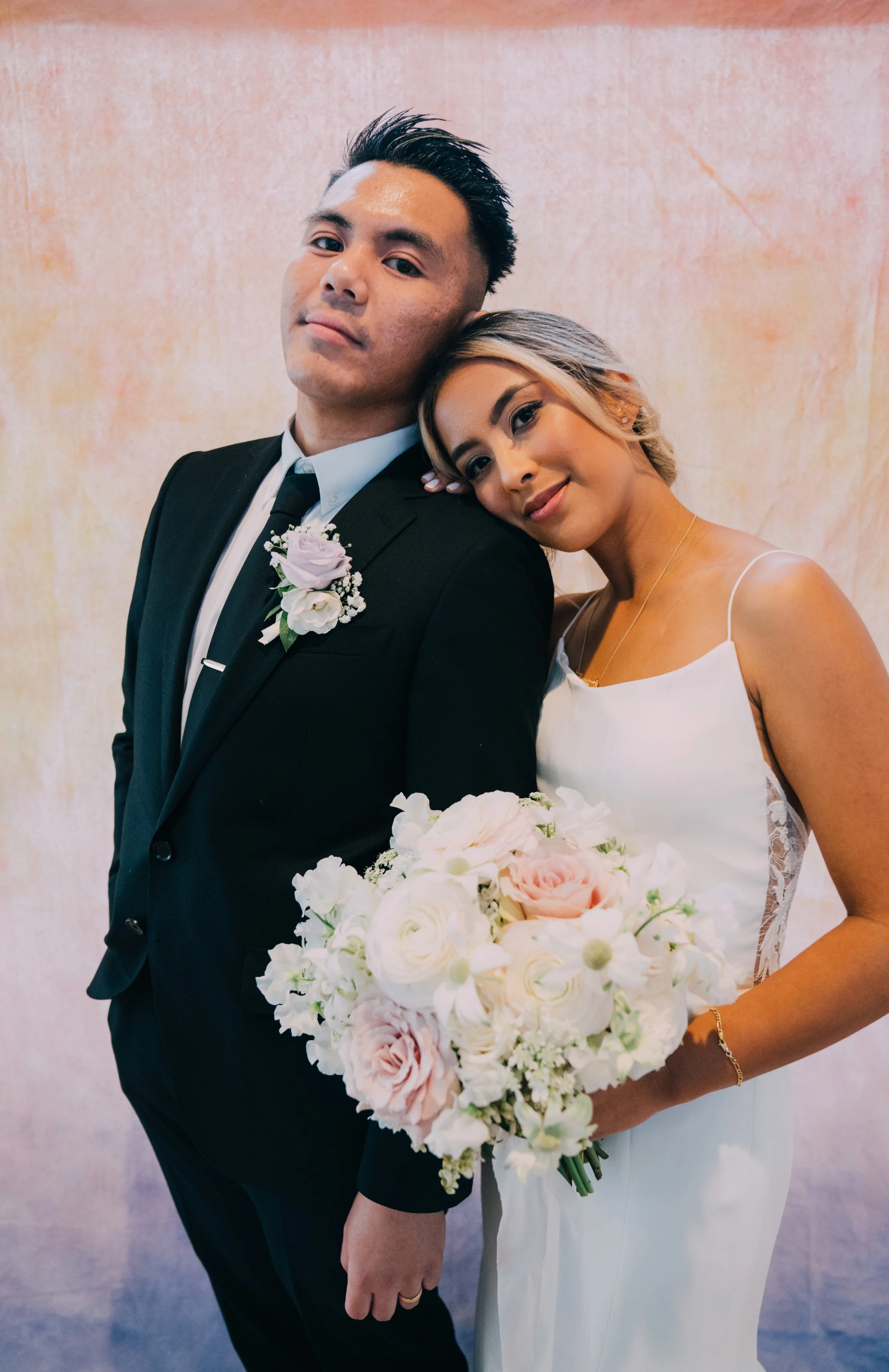 A bride and groom pose together, with the bride resting her head on the groom's shoulder. The groom is in a black suit with a white shirt, black tie, and a boutonniere. The bride wears a white dress with thin straps and holds a large bouquet of pale 