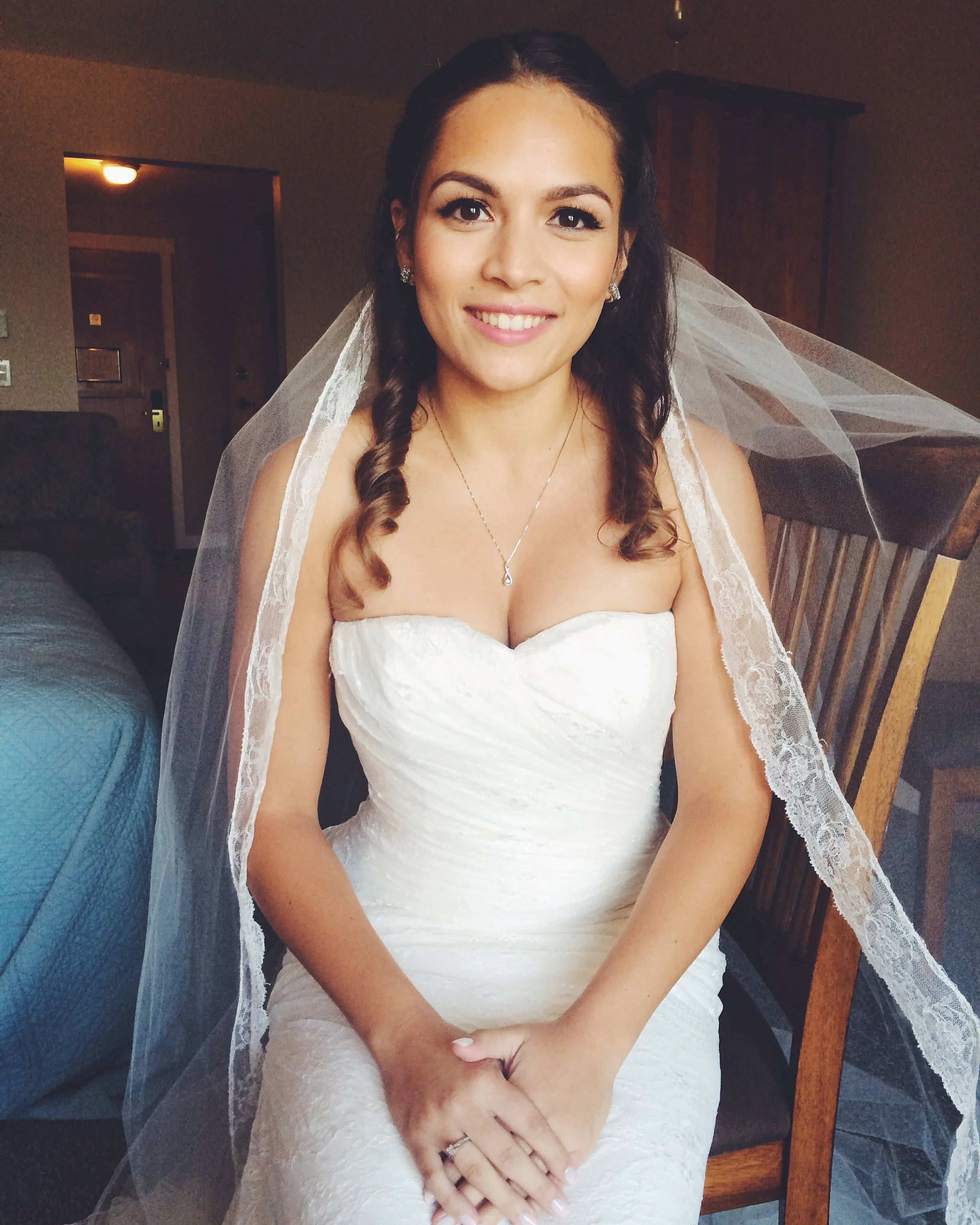 A woman in a white wedding dress with lace details, wearing a veil, earrings, and a necklace, smiling with her hands clasped in front of her, sitting indoors.