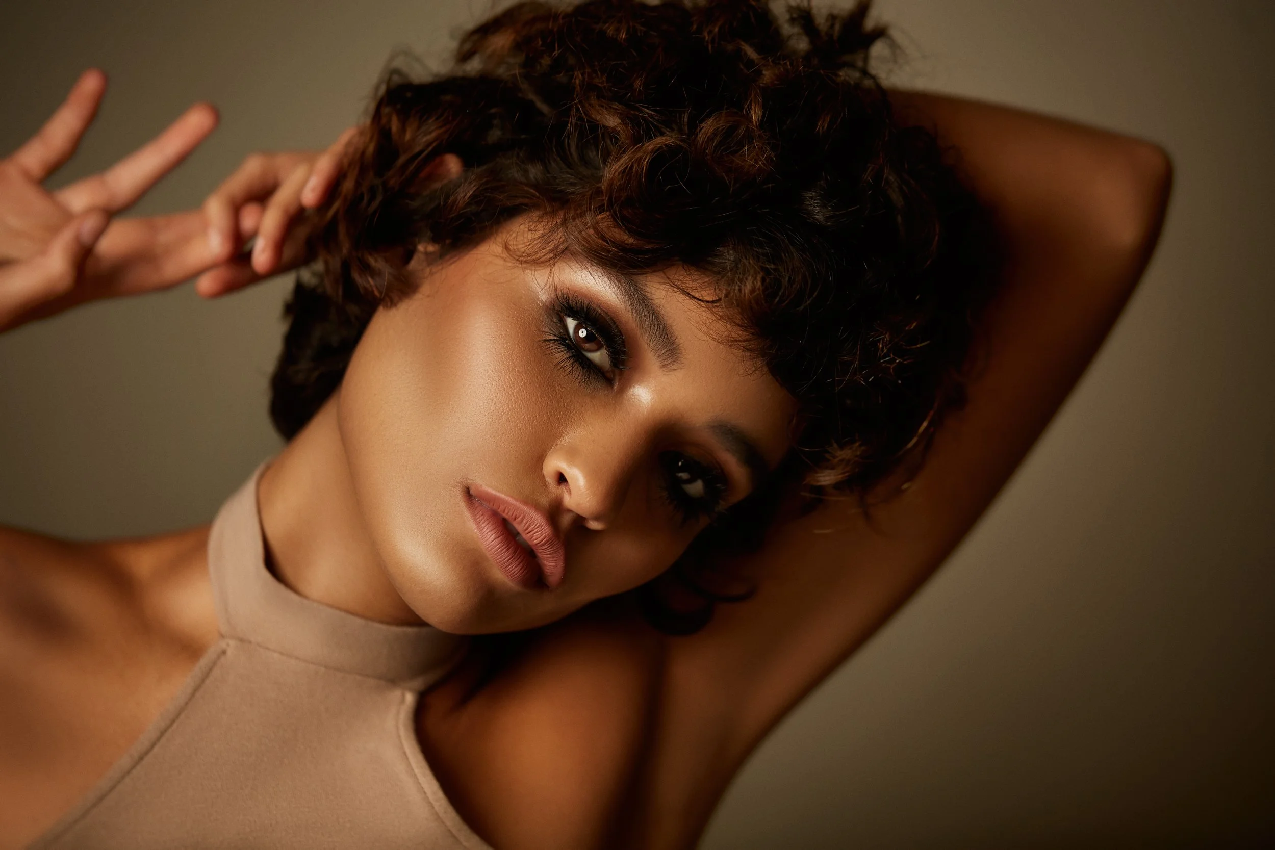 A woman with short, curly brown hair and smoky eye makeup posing with one arm raised behind her head.