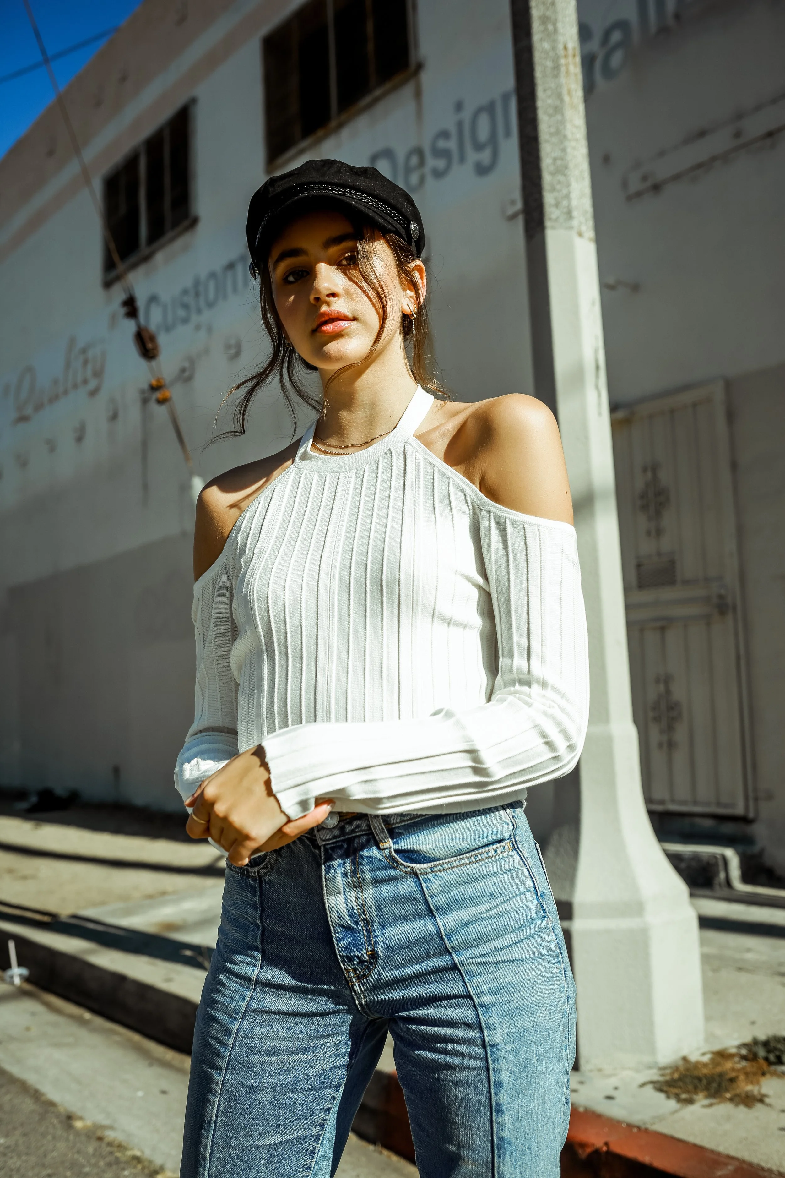 Young woman with dark hair, wearing a black cap and a white off-shoulder sweater, standing outdoors in front of a building with a sign that reads 'Quality Custom Design'. She has a neutral expression and is looking at the camera.
