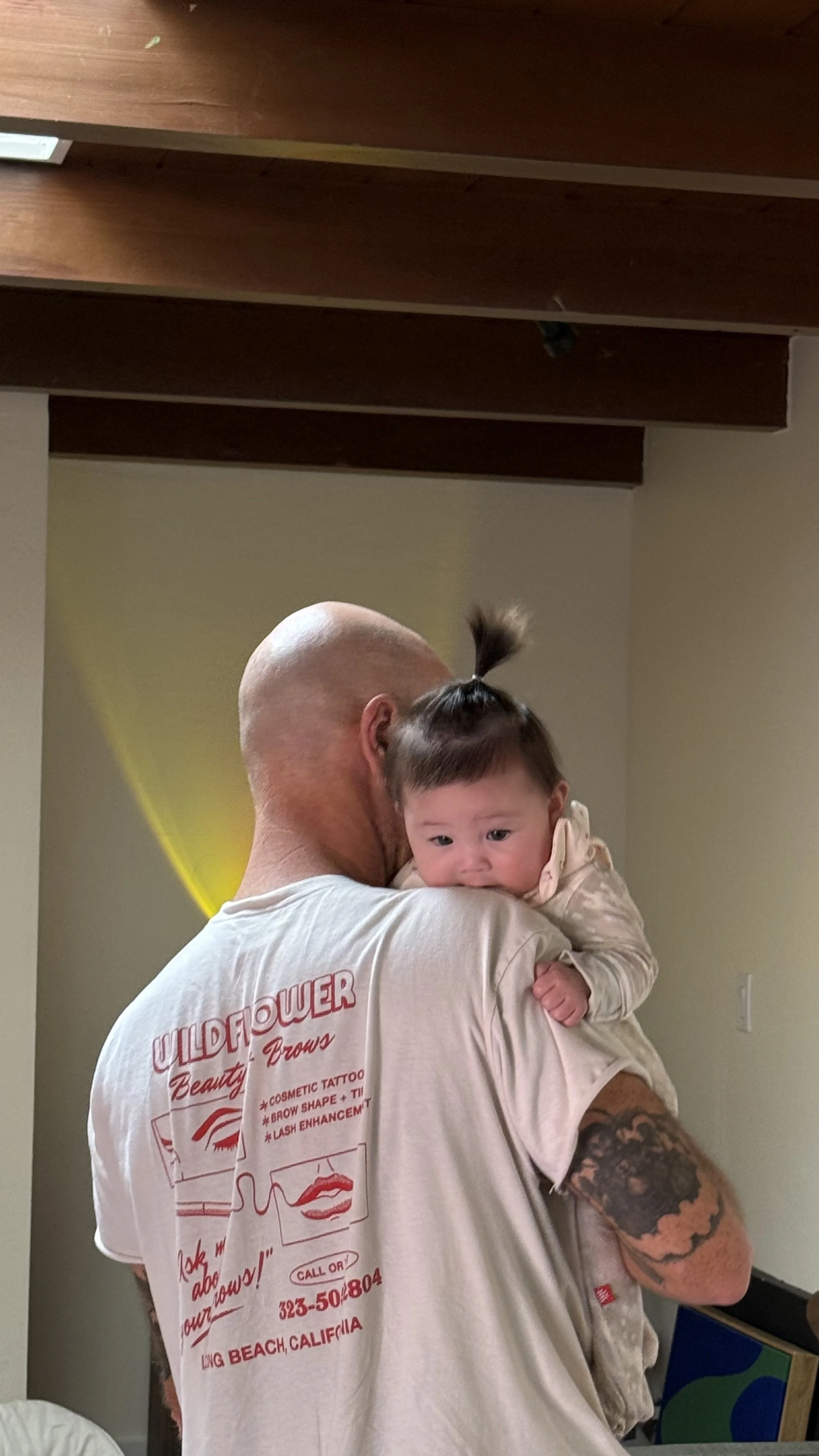 A man holding a baby in his arms. The man is wearing a light-colored Wildflower Beauty + Brows merch shirt with red print on the back. The background has a wall and a ceiling with wooden beams.
