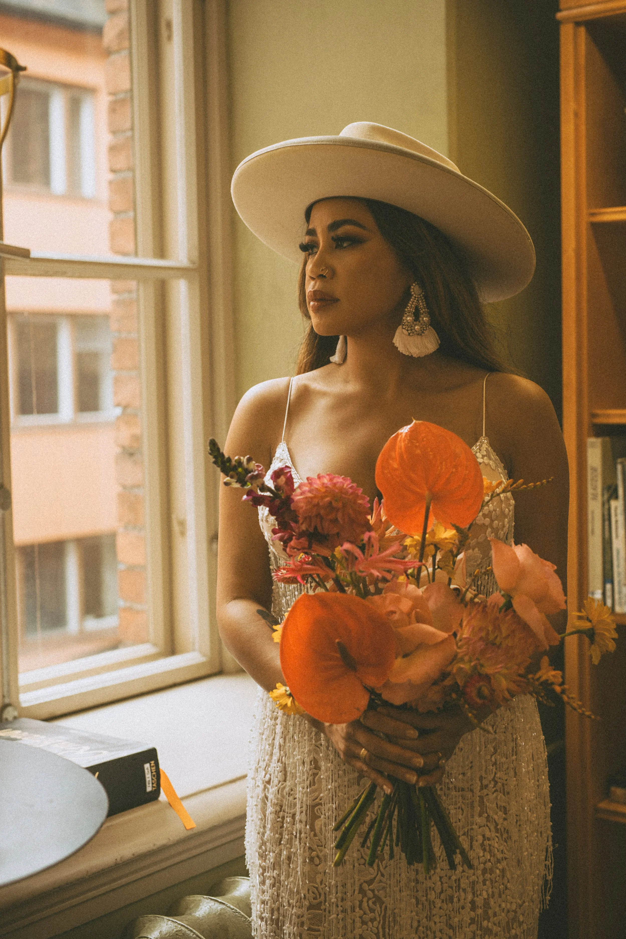 A woman in a white dress and wide-brimmed hat holding a bouquet of flowers, standing indoors near a window with a brick building visible outside.