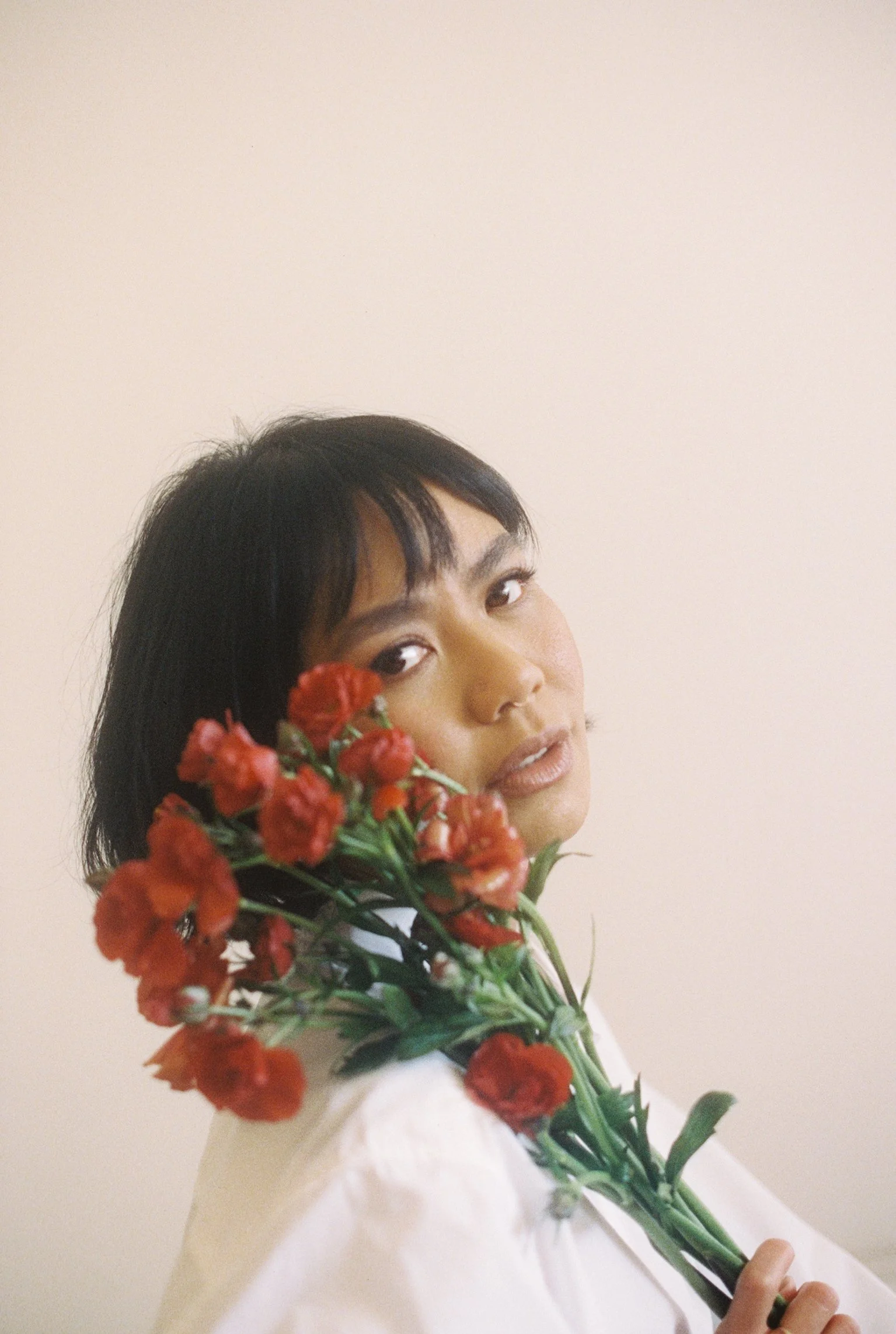 Rachelle holding a large bouquet of red flowers in front of her face, partially obscuring her features, against a plain beige background.